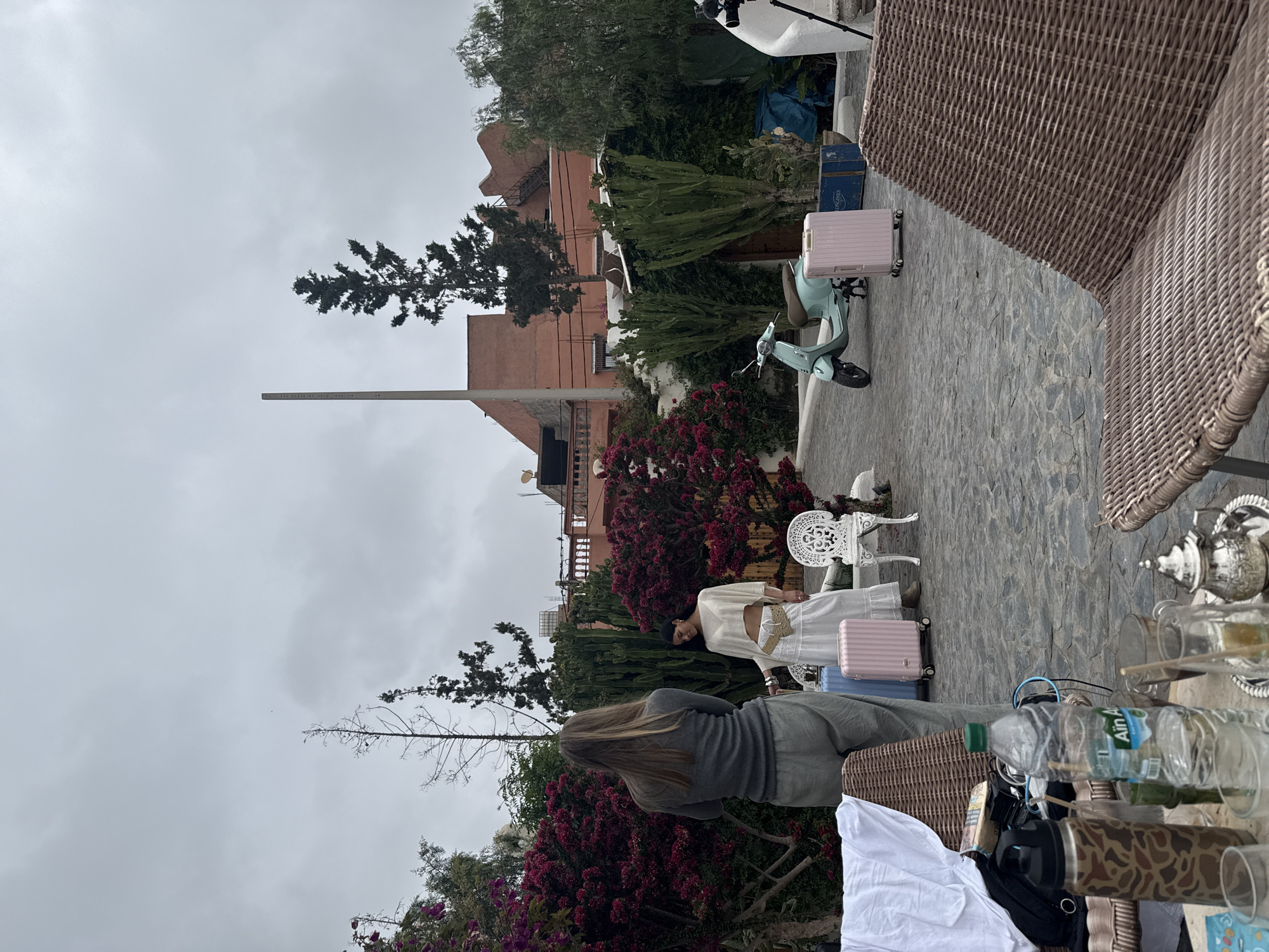 A medium shot shows a scene at Lalla Takarkoust, Morocco, under an overcast sky. A light green scooter and luggage sit on a stone patio. Two women stand amongst flowering bushes. A white metal chair and a tea set are on the patio. Various water bottles and glasses are on a table. In the background are buildings with a unique architectural style and a communications tower.