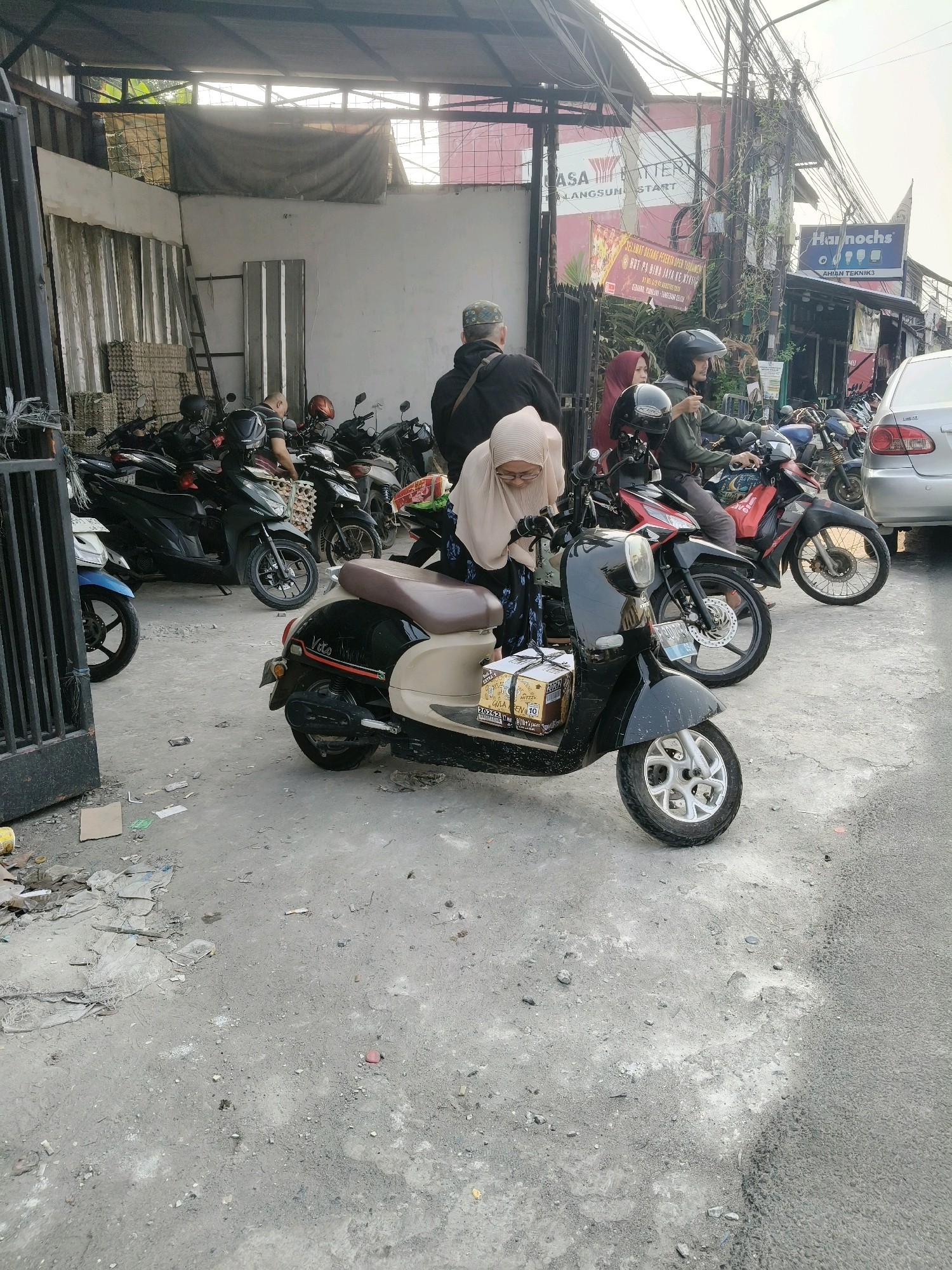 The image depicts an outdoor scene likely in South Tangerang, Indonesia, featuring a row of parked motorcycles. In the foreground, a black and beige scooter is prominently displayed, with a person wearing a beige hijab and glasses seated on it, looking down at a package secured on the front. To the left of the scooter, a blue motorcycle is visible. Behind these, numerous other motorcycles are parked, some with riders or people interacting with them. A man wearing a black jacket and a patterned cap is standing near some of the motorcycles. In the background, buildings with signage, including "Harnochs" and "ASA BATTERY," are visible, along with overhead wires and some foliage. The ground is paved with concrete and appears somewhat unkempt, with scattered debris. The lighting suggests it is daytime, possibly late afternoon.
