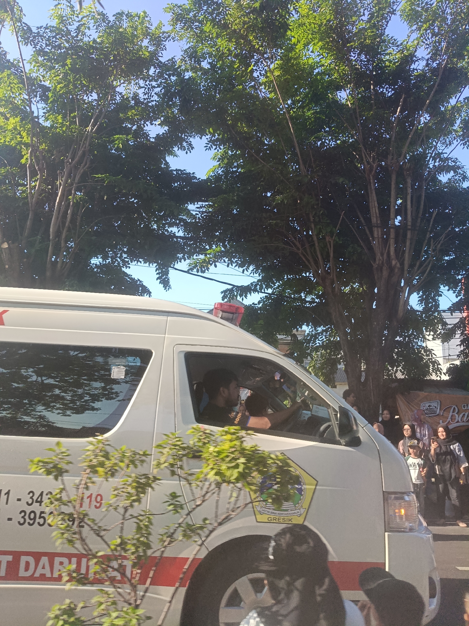 An ambulance with the words "DARURAT" and what appears to be a phone number and the text "GRESIK" visible on its side is driving down a street lined with trees and buildings. A man is visible driving the ambulance. In the background, several people are gathered on the sidewalk, some looking towards the ambulance. The scene is illuminated by sunlight, suggesting it is daytime.
