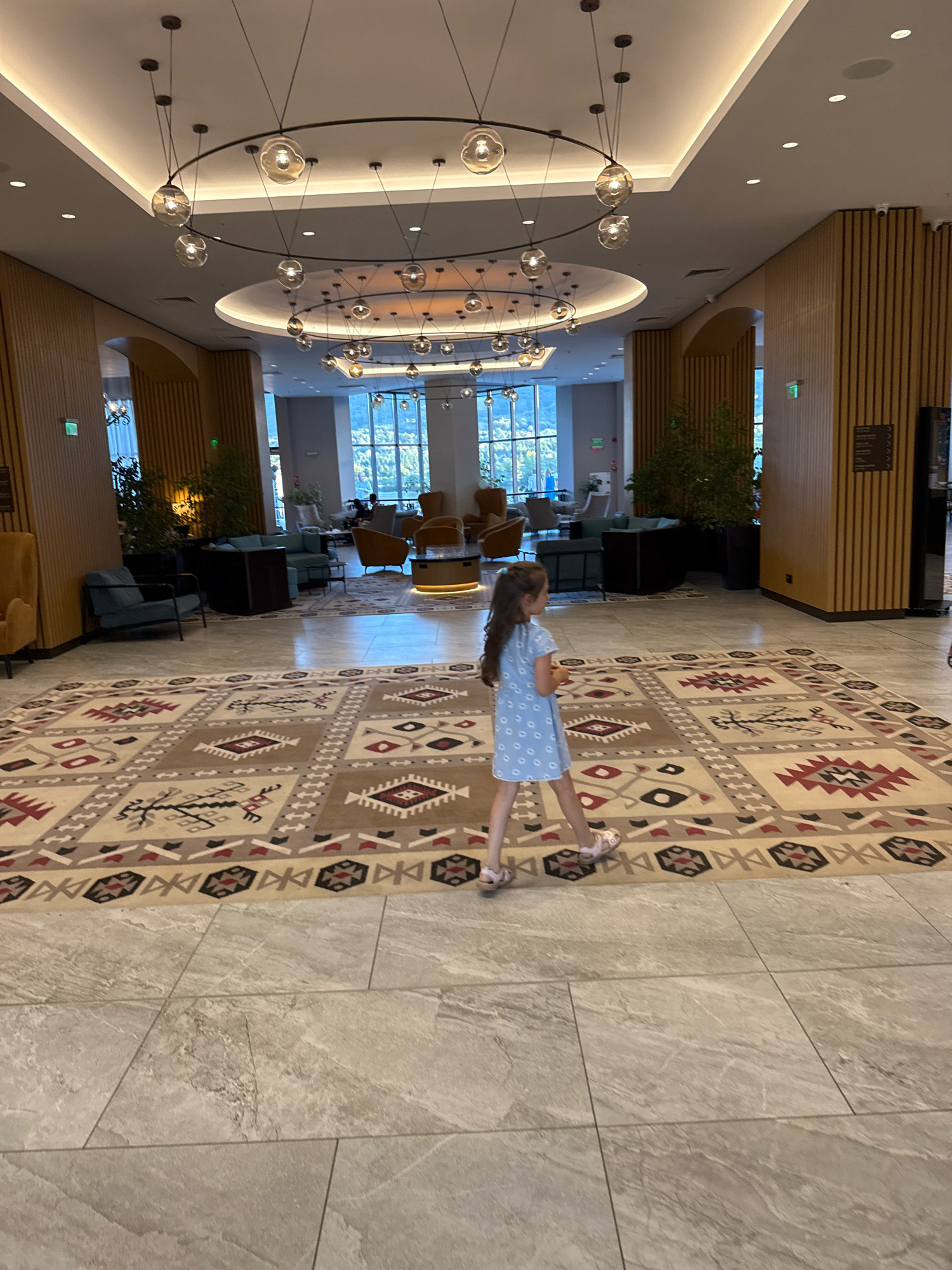 A young girl in a light blue dress and pink sandals walks across a large, ornate rug in a modern, well-lit hotel lobby. The rug features geometric patterns in beige, brown, red, and black. The lobby has a high ceiling with a circular lighting fixture and recessed lighting. In the background, there are seating areas with upholstered chairs and sofas, as well as large windows offering a view of a natural landscape. The floor is tiled with large, light-colored tiles. The overall impression is one of a contemporary and spacious hotel. This image was taken in the city of Razliv, Bulgaria.
