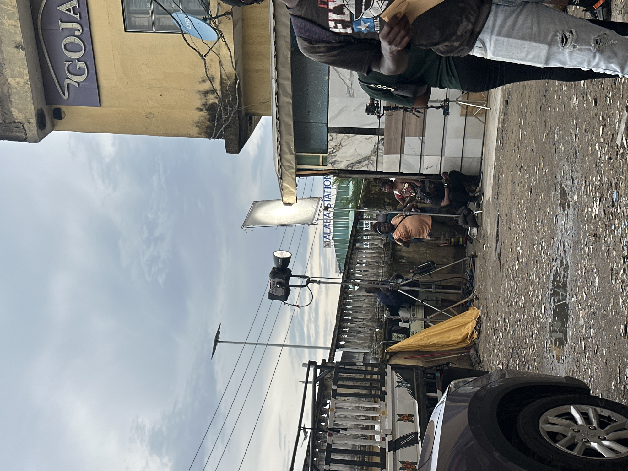 The image depicts a film set in a bustling area of Lagos, Nigeria, near Alaba Station. A film crew is actively engaged with cameras, lighting equipment (including a large reflector), and other accessories. Several individuals are visible in the frame, some operating equipment and others likely acting. The environment consists of a partially wet street littered with debris, suggesting recent rain. Buildings are visible in the background, with one displaying a "GOJA" sign. A car is parked on the side of the street, partially obstructing the view. The overall scene conveys the active nature of film production in an urban setting.