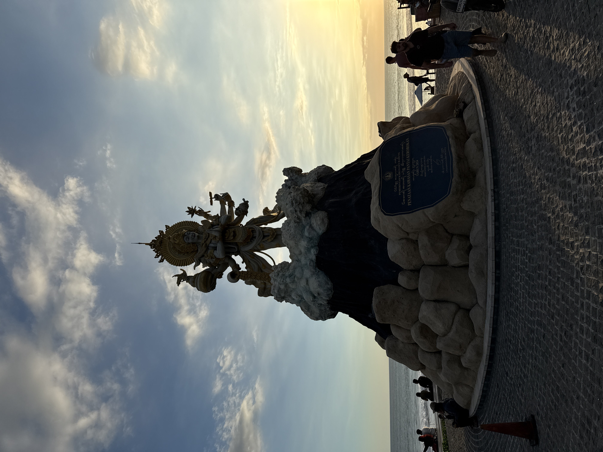 The image shows a statue of a deity in North Kuta, Indonesia. The statue has many arms and intricate detailing. It sits atop a dark, rocky structure that is surrounded by lighter-colored stones. A plaque is attached to the structure. The sky behind the statue is a mix of blue and cloudy white, with a hint of yellow near the horizon, suggesting a sunset or sunrise.
Along the edge of the paved area, there are people standing and walking near the water. A motorcycle is parked nearby. The pavement is made of dark, small tiles. An orange traffic cone sits at the bottom of the frame. The OCR text on the plaque mentions "PEMERINTAH KABUPATEN BADUNG" (Government of Badung Regency) and "PENATAAN KAWASAN PANTAI KEROBOKAN" (Arrangement of the Kerobokan Beach Area), indicating the location is Kerobokan Beach. It also mentions the year 2024.