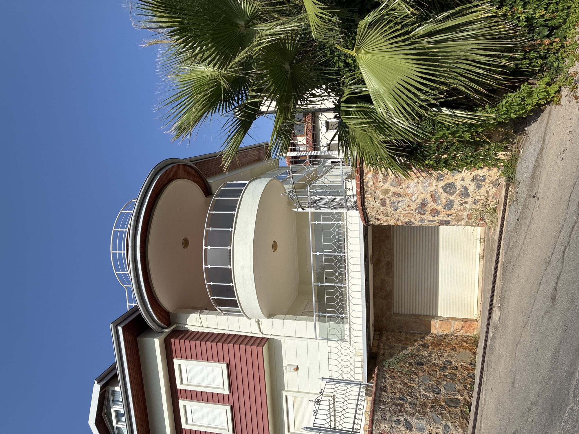 The image captures a portion of a multi-story building in Alanya, Turkey, under a clear blue sky. The building features a prominent curved balcony with a white railing and tinted glass panels. The exterior walls are painted a light cream color, with a section of dark red horizontal siding. A sloped roof with red trim is visible at the top. In front of the building, a large palm tree with vibrant green fronds partially obscures the view. The building is situated on a sloped lot, with a stone-clad retaining wall supporting the elevated structure. A cream-colored roll-up garage door is visible at the base of the building. A metal decorative fence runs along the edge of the elevated area. The foreground shows a portion of an asphalt road.
