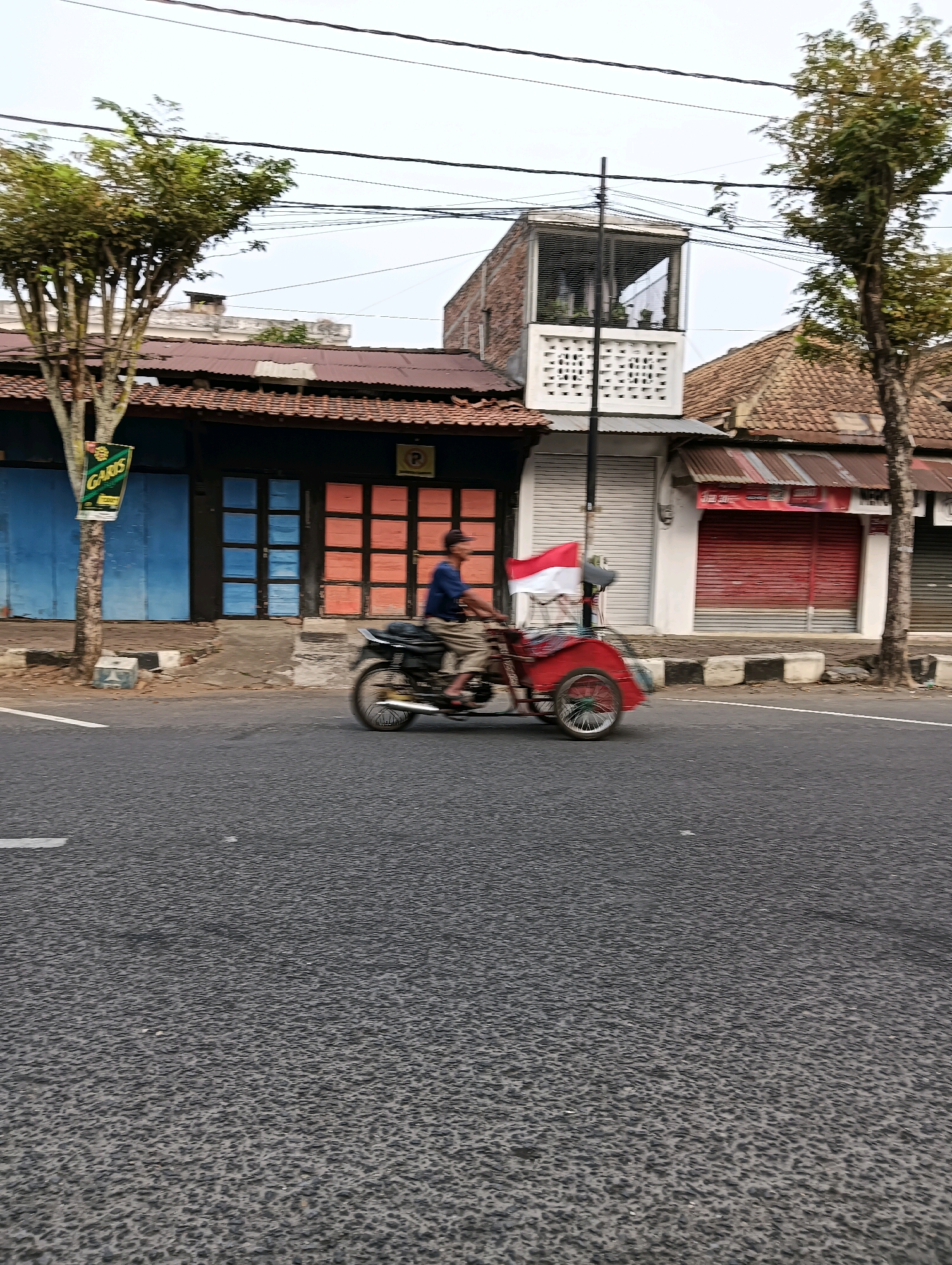 A red and white Indonesian flag flies from a red pedicab with a single rider on a street in Purworejo, Indonesia. The pedicab is moving from left to right across the frame, and the rider is wearing a blue shirt and light-colored pants. The street is paved and appears to be wet or reflective. In the background, there are several storefronts with metal roll-up doors, some painted blue and some red. Trees with green foliage are visible on the left and right sides of the frame. Overhead, power lines are strung across the sky.