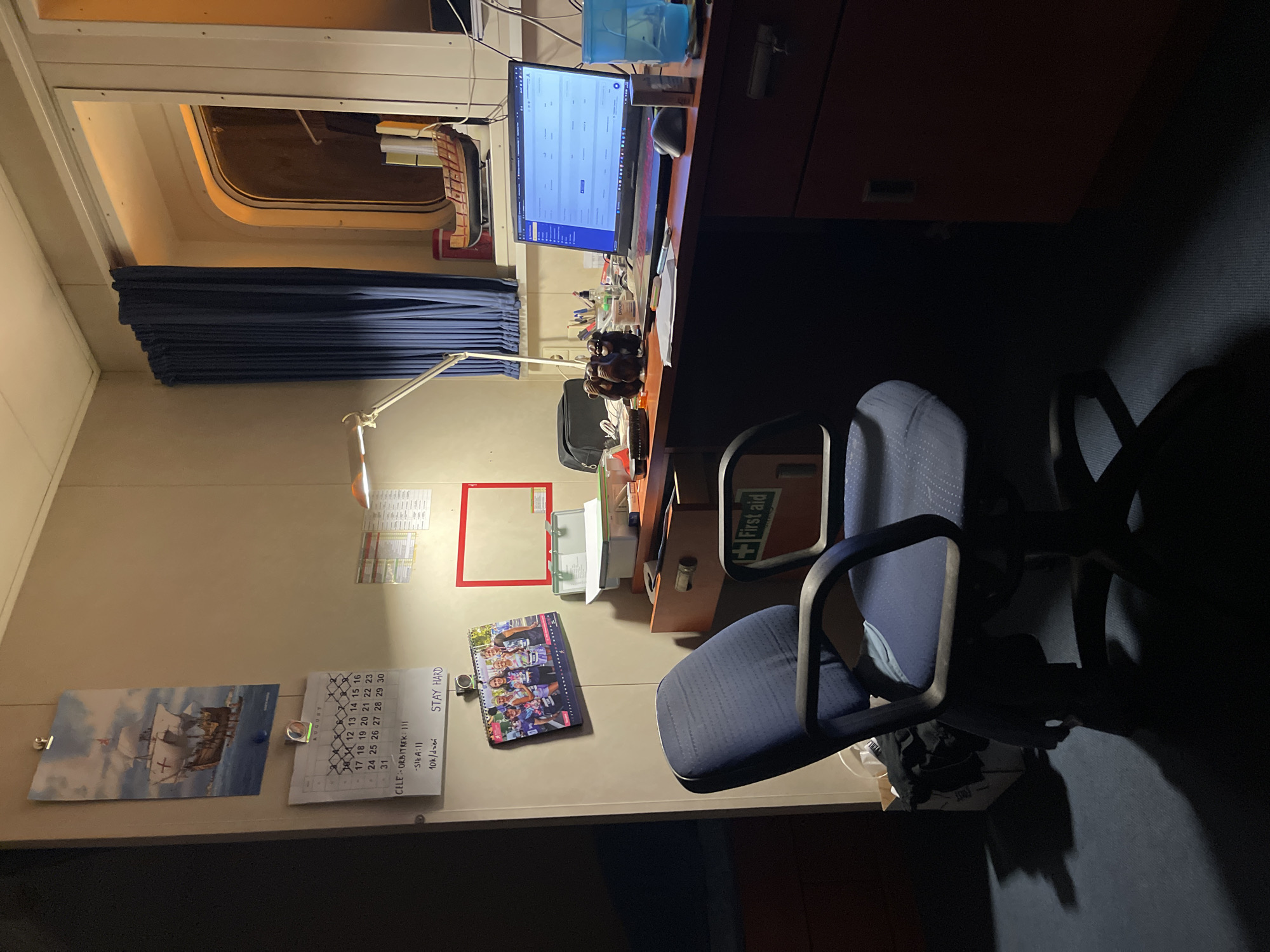 This image depicts an indoor scene, likely a personal workspace or cabin, with no discernible connection to Sassako-Bégini, Ivory Coast. The environment is dimly lit, with the primary light source appearing to be a desk lamp.
In the foreground, a dark blue office chair with a patterned fabric is positioned in front of a wooden desk. To the left of the chair, a portion of a dark cabinet or furniture is visible.
The desk itself is cluttered with various items. A laptop, open and displaying a screen with a table-like interface, is the most prominent object on the desk. To the left of the laptop are a mouse, a pen, several small decorative figures resembling monkeys, a black bag, and a few other unidentifiable objects. Further to the left, a white box or container with some contents is visible. Behind the laptop, a model ship is displayed on a shelf or surface.
On the wall behind the desk, several items are mounted. A calendar showing the month of August is visible, with dates marked and some handwritten text below. Above the calendar, a picture of a sailing ship is displayed. To the right of the calendar, a red-framed memo board or display area is present.
To the right of the desk, a blue curtain is drawn across a window. The window itself is visible, appearing to be a porthole-style window with internal lighting. On the far right edge of the image, a portion of a red cabinet or drawer unit is visible. A green box labeled "First aid" is affixed to the side of the desk or a nearby surface, visible to the right of the chair.