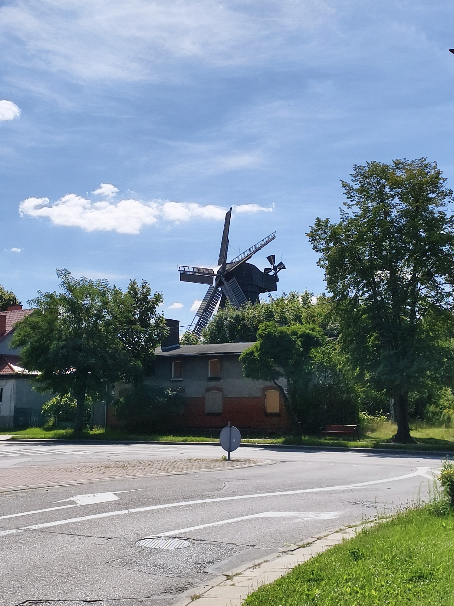 The image depicts a Dutch windmill situated behind a residential building and surrounded by trees. The windmill is made of dark wood with four blades, one of which is partially obscured. The building appears to be a two-story structure with a dark roof and brick accents. The windmill and building are located in a semi-urban environment, as evidenced by the presence of a road, crosswalk, and street signs. A large, leafy tree is prominent in the foreground, to the right of the windmill. The sky is blue with scattered white clouds. The scene suggests a sunny day. This windmill is known as the "Wiatrak" in Tczew, Poland, and is a historical landmark in the city.