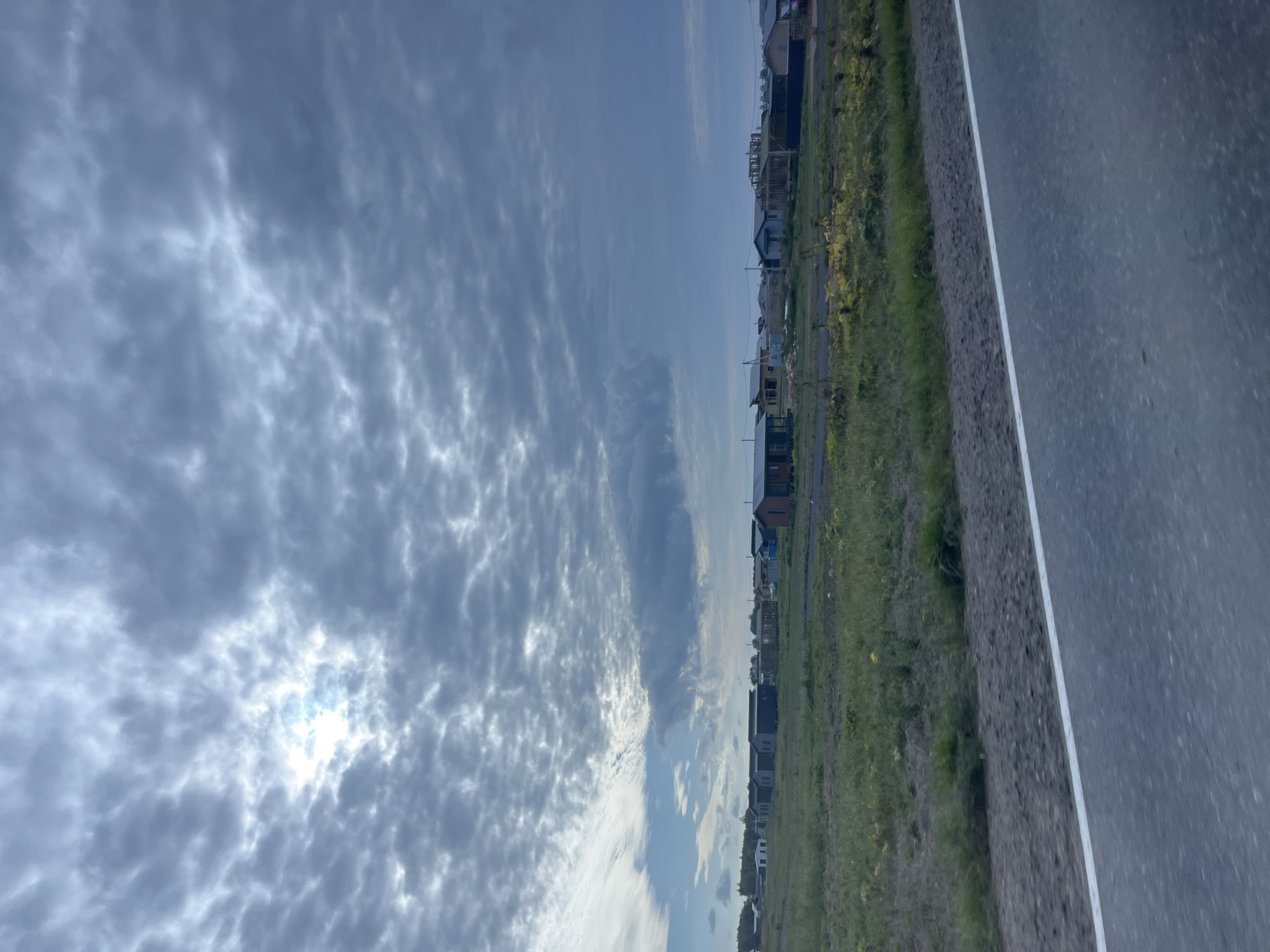 The image depicts a cloudy sky over a rural landscape in the vicinity of Glyadino, Russia. A partially obscured sun peeks through the dense, grey clouds, casting a diffused light. Below the sky, a field of dry grass and some scattered yellow wildflowers stretches towards a cluster of houses and unfinished structures in the distance. The houses appear to be single-story, with various roof styles and some under construction. A paved road with a white line marking its edge runs diagonally across the foreground, suggesting a roadside perspective. The overall impression is one of a developing rural community on an overcast day.