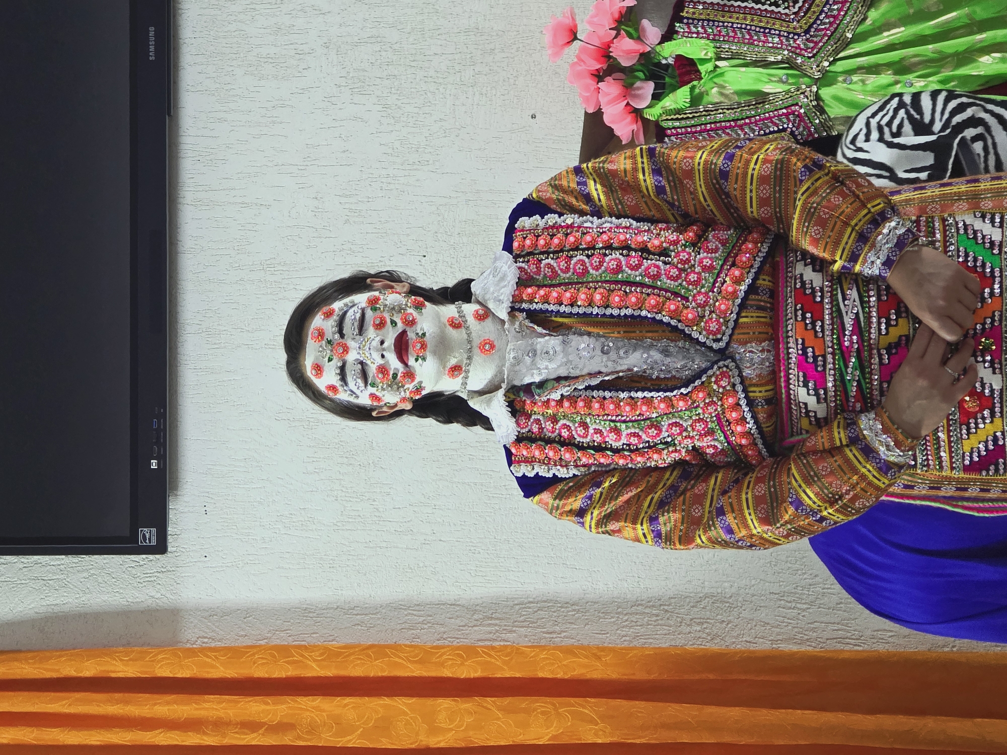 A person with their eyes closed and face painted white with red floral embellishments stands in front of a textured white wall. They wear a brightly colored, ornate traditional garment adorned with numerous small pink and red beads and sequins. The garment features intricate patterns and embroidery. To the left of the person is an orange curtain with a floral pattern. To the right, a bouquet of pink flowers sits next to another green traditional garment. A black television is mounted on the wall above the person. The scene appears to be indoors, likely in Ribnovo, Bulgaria, a village known for its preserved traditional Bulgarian wedding customs.