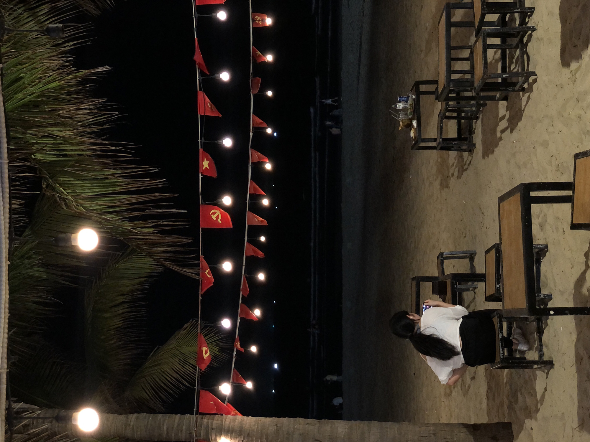 This media file shows a nighttime scene on a sandy beach in Da Nang, Vietnam. The foreground features palm fronds and a string of red flags interspersed with glowing lights, suggesting a festive or celebratory atmosphere. In the background, several people are visible, some walking along the shoreline while others are seated at tables. One person, a young woman with her hair tied in a ponytail, is seated at a table and appears to be taking a photo with her phone. She is wearing a white top and black pants.
The setting is a beachside establishment, likely a restaurant or cafe, with simple wooden tables and chairs set up on the sand. The lighting from the string lights and a few overhead lamps casts shadows on the sand, contributing to the nighttime ambiance. The time of day is clearly nighttime, and the weather appears to be clear, given the visibility of the stars faintly in the distance and the people being out on the beach.
The red flags with yellow stars are a prominent visual element and are indicative of Vietnam. This, combined with the location context provided, strongly suggests the scene is in Vietnam. There is no visible text in the image. The overall mood seems relaxed and enjoyable, typical of a beach evening outing.