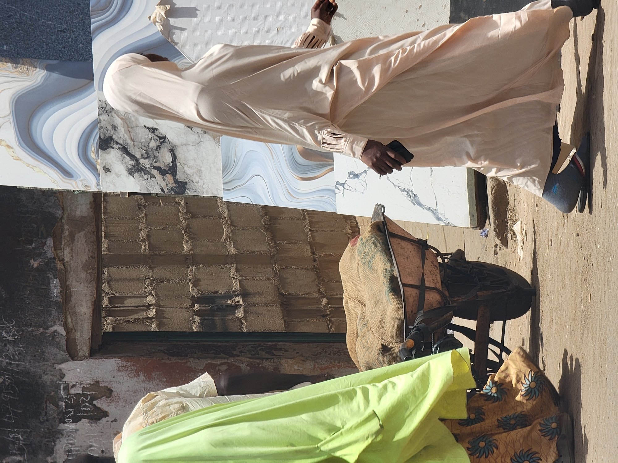 An outdoor scene in Maiburiji, Nigeria, presents a man wearing a long, light-colored robe. He stands on a street next to a wall, alongside several stacked decorative tile pieces. He is holding a small, dark object, likely a mobile phone, in his right hand. Adjacent to him is a bicycle with a padded seat and bags loaded onto the rear. A green cloth is partially visible near the bottom of the frame. The lighting suggests it is daytime.