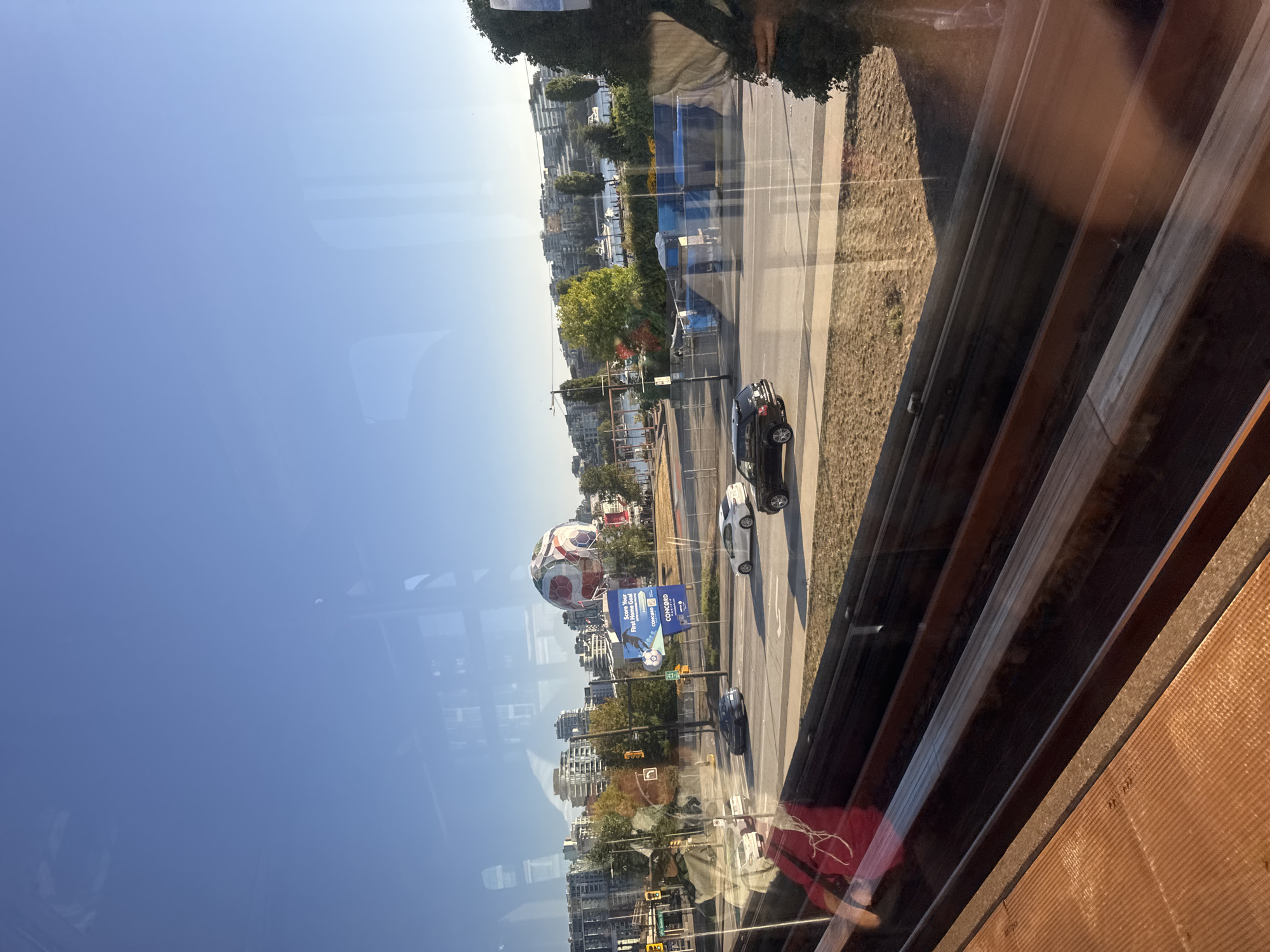 This image captures a view through a window, likely from a moving vehicle or building, showcasing a cityscape with a clear blue sky. In the mid-ground, a large, inflatable soccer ball structure is prominently displayed, seemingly part of an advertisement or event. Adjacent to it are billboards, one of which reads "Score Your First Home Goal" and features the "Concord Pacific" logo, indicating a real estate development or promotion.
The scene includes a street with several vehicles, including a black SUV, a white sedan, and a dark-colored sedan, suggesting moderate traffic. Buildings of various heights, characteristic of a city, line the background, with some appearing to be residential complexes and others commercial structures. Trees with autumn foliage add color to the urban landscape. Further in the distance, a body of water with boats docked is visible, and beyond that, more buildings and a distant horizon. The foreground shows the interior of the structure from which the photo is taken, with what appears to be wooden seating or track elements. The reflection of a person in a red shirt is visible on the window in the lower left quadrant of the image. The overall impression is a bright, sunny day in a developed urban area. The presence of "Concord Pacific" and the architectural style of the buildings strongly suggest this location is Vancouver, Canada, likely in the False Creek area.