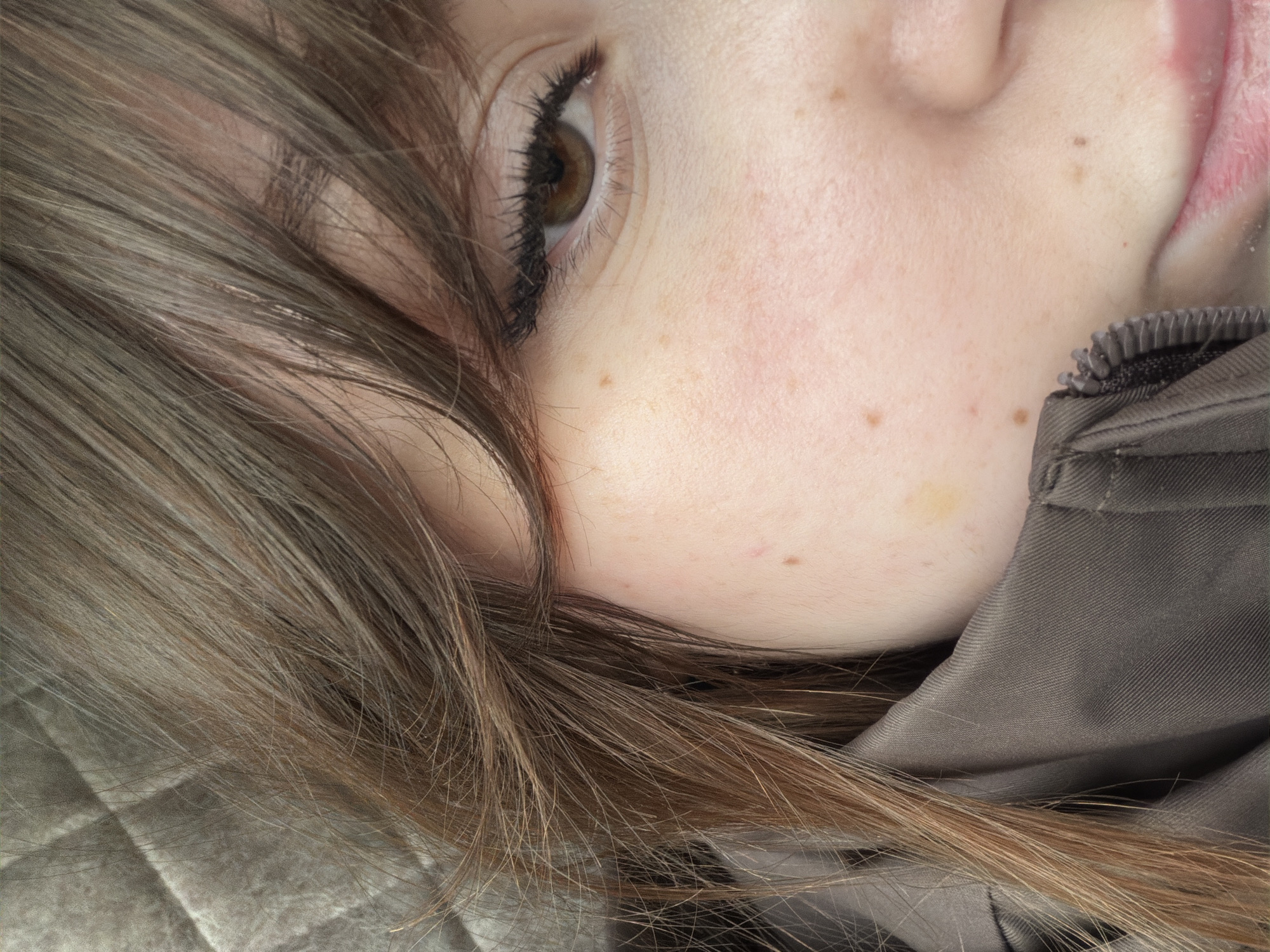 A close-up, tilted view captures the right side of a person's face. The subject has fair skin, brown freckles, and brown hair with lighter highlights. The hair partially covers their face. Their right eye is visible, showcasing dark lashes. The person's cheek rests against a dark gray fabric, possibly clothing. The image lacks any visible contextual clues to pinpoint a location in Chaikovsky, Russia. No specific actions or interactions are discernible. The environment is limited to the immediate surroundings of the subject's face and clothing.