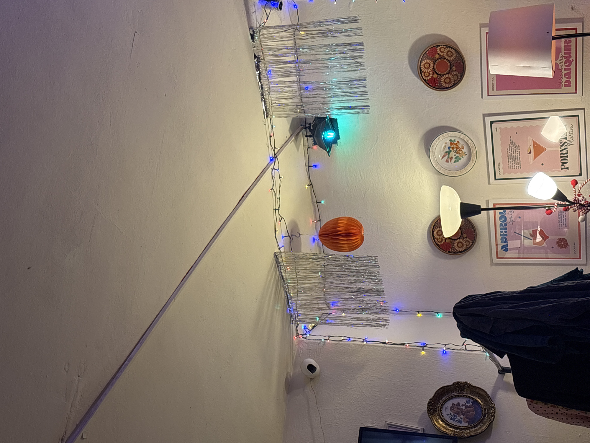 The image shows an interior setting with a white wall adorned with various decorative elements. There are framed cocktail recipe posters for "Strawberry Daiquiri," "Pornstar Martini," and "Aperol Spritz." Several decorative plates are also mounted on the wall. Strings of multi-colored lights and silver tinsel hang from the ceiling. An orange paper lantern is suspended from the ceiling. A surveillance camera is mounted on the wall. A black garment hangs from a rack, partially obscuring the lower right area. A television is visible in the lower center of the image. The location appears to be a decorated interior space. The city and country of Belgrade, Serbia, are not evident from the image.