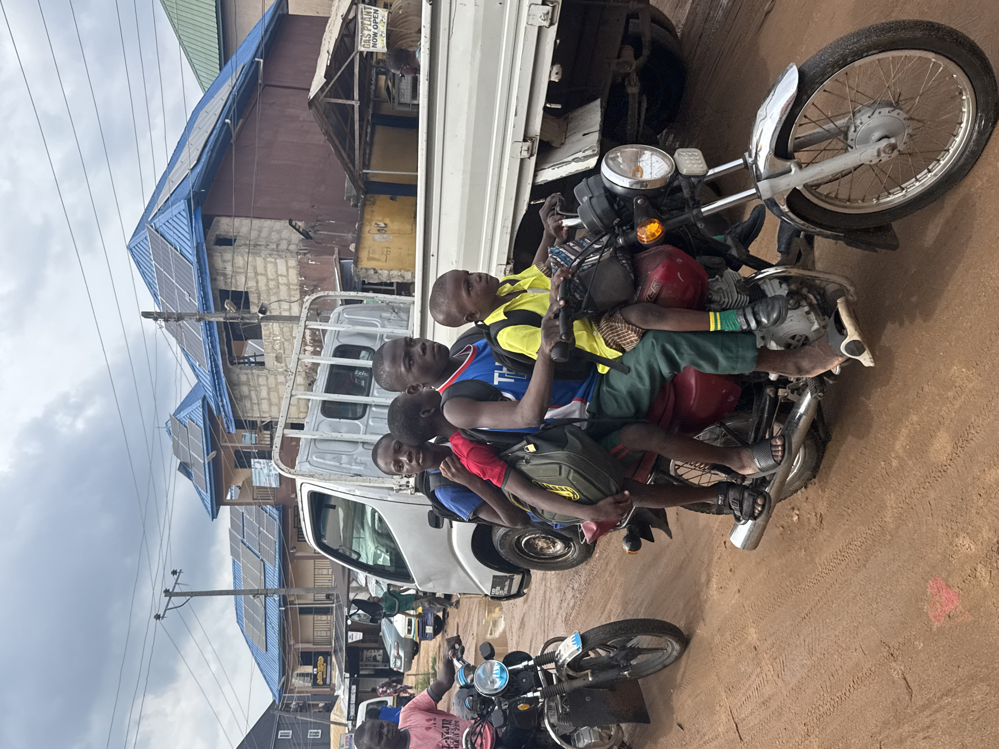 The image depicts a vibrant street scene in Ihiagwa, Nigeria, under an overcast sky. The primary subjects are a group of five young males, appearing to be children and teenagers, overloaded on a red motorcycle in the middle of a dusty, unpaved road. The youngest child, wearing a yellow shirt, sits near the handlebars, while the others are packed closely behind him, one in a blue and black top and another in a green and black outfit. Most are wearing sandals or flip-flops.
In the foreground, another man is seated on a separate black motorcycle, facing away from the camera. A white utility truck is visible behind the crowded motorcycle, with "Ezio" written on its side. The backdrop consists of several buildings, some under construction or in a state of disrepair, featuring concrete block walls and blue corrugated metal roofs. Notably, multiple solar panels are installed on the rooftops of these buildings, indicating a reliance on renewable energy. Overhead, numerous power lines stretch across the cloudy sky.
A partially visible sign in the background reads "GAS PLANT NOW OPEN." The scene captures a snapshot of daily life in a community where motorcycles are a common mode of transport, often carrying multiple passengers, and local infrastructure includes both traditional construction and modern renewable energy solutions.