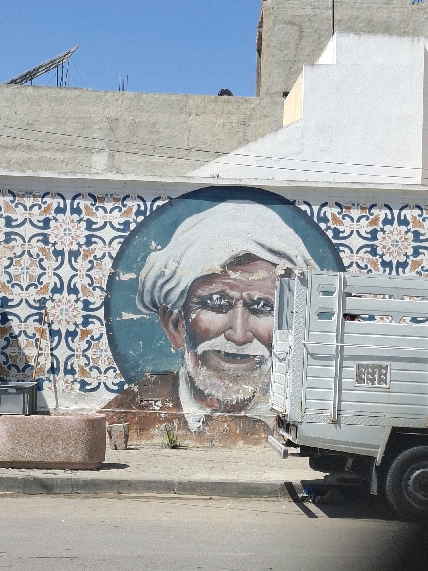 The image depicts a mural of an elderly man with a white turban and beard painted on a wall in Bennane, Tunisia. The mural is set against a patterned background of blue and orange geometric shapes. To the right of the mural, a white truck is parked, partially obscuring the painting. In the foreground, a concrete curb and a street are visible. The sky above is clear and blue.