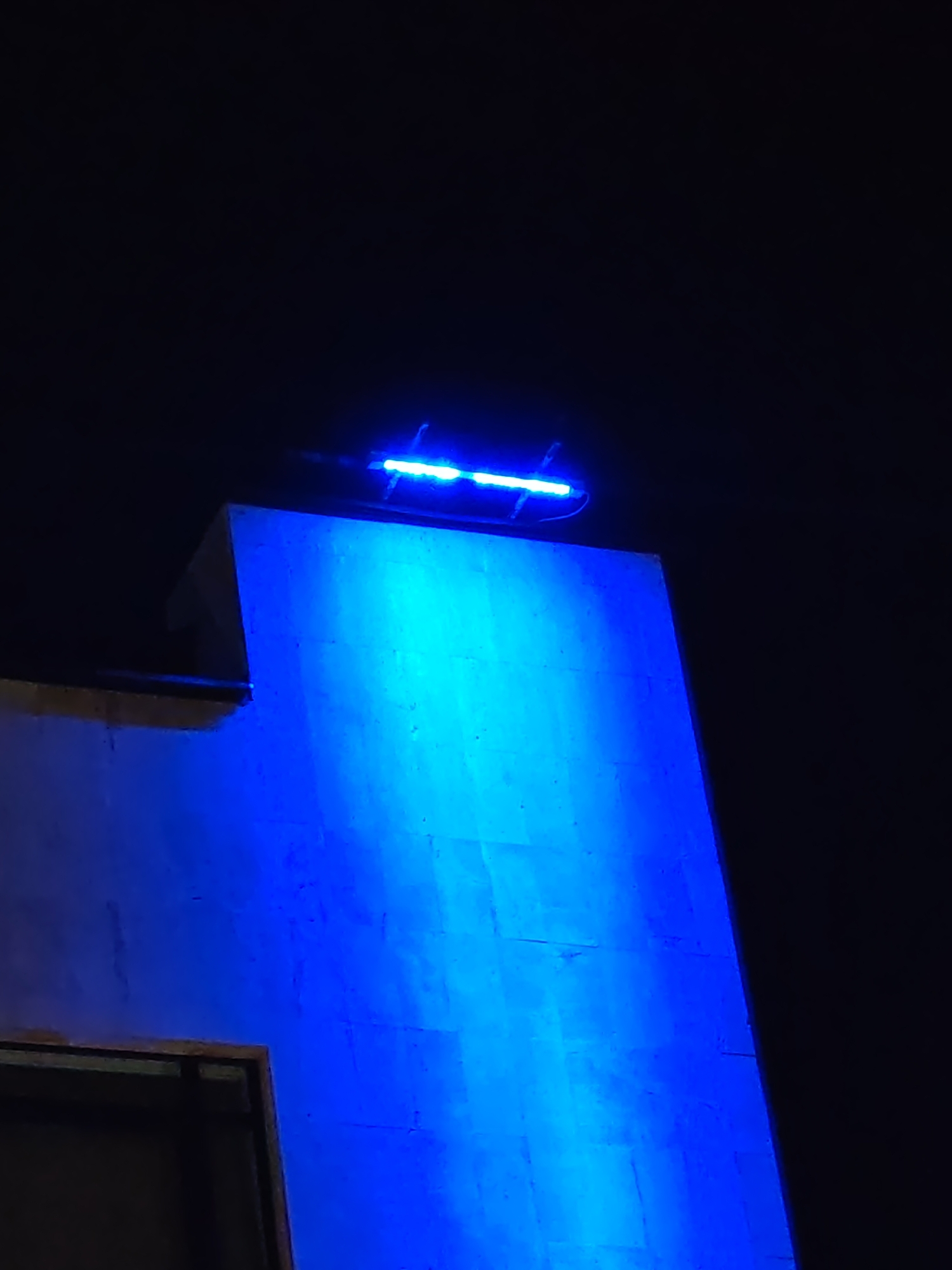 The image captures a nighttime scene in Veliki Preslav, Bulgaria, featuring a large, vertically oriented architectural structure. A linear light fixture, emitting a strong blue light, is mounted horizontally across the top edge of this structure. The blue light casts a vibrant illumination down the facade, highlighting the wall's texture, which appears to be composed of stone or concrete blocks with visible seams. To the upper left, a darker, stepped architectural element is partially visible, receding into shadow. In the lower-left foreground, a dark rectangular opening with a visible brown frame is present. The sky and surrounding areas are uniformly dark, indicating an absence of ambient light beyond the blue illumination. No individuals or other specific objects are discernible.