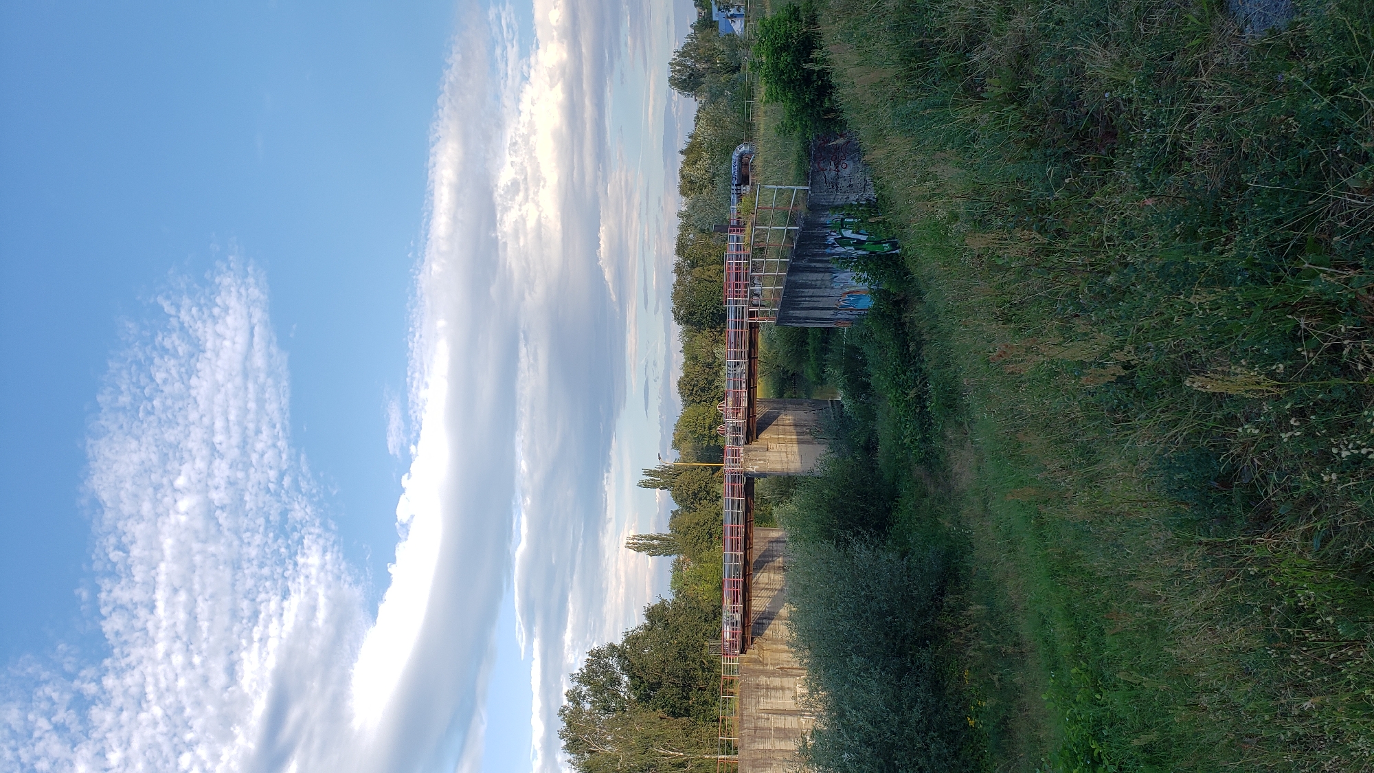 The image captures a view of a bridge over a canal or river, situated in the city of Prešov, Slovakia. The bridge features a red and white railing. Concrete abutments support the bridge, and one of these abutments has graffiti on it. The scene is framed by lush green vegetation, including trees and tall grass, with a cloudy blue sky overhead. The overall impression is one of a somewhat overgrown, possibly industrial or semi-urban landscape.