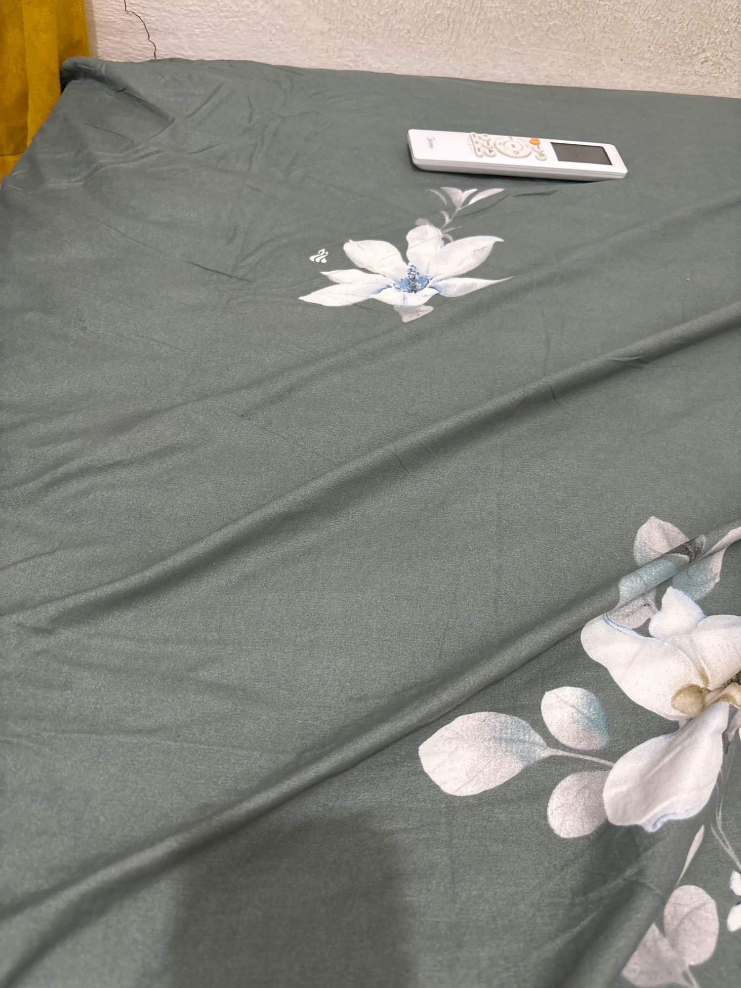 A white remote control is placed on a dark green duvet cover with a floral pattern. The duvet cover is slightly wrinkled, and the remote control is positioned horizontally. The background shows a textured wall and a glimpse of a yellow curtain. The scene appears to be an indoor setting, possibly a bedroom.