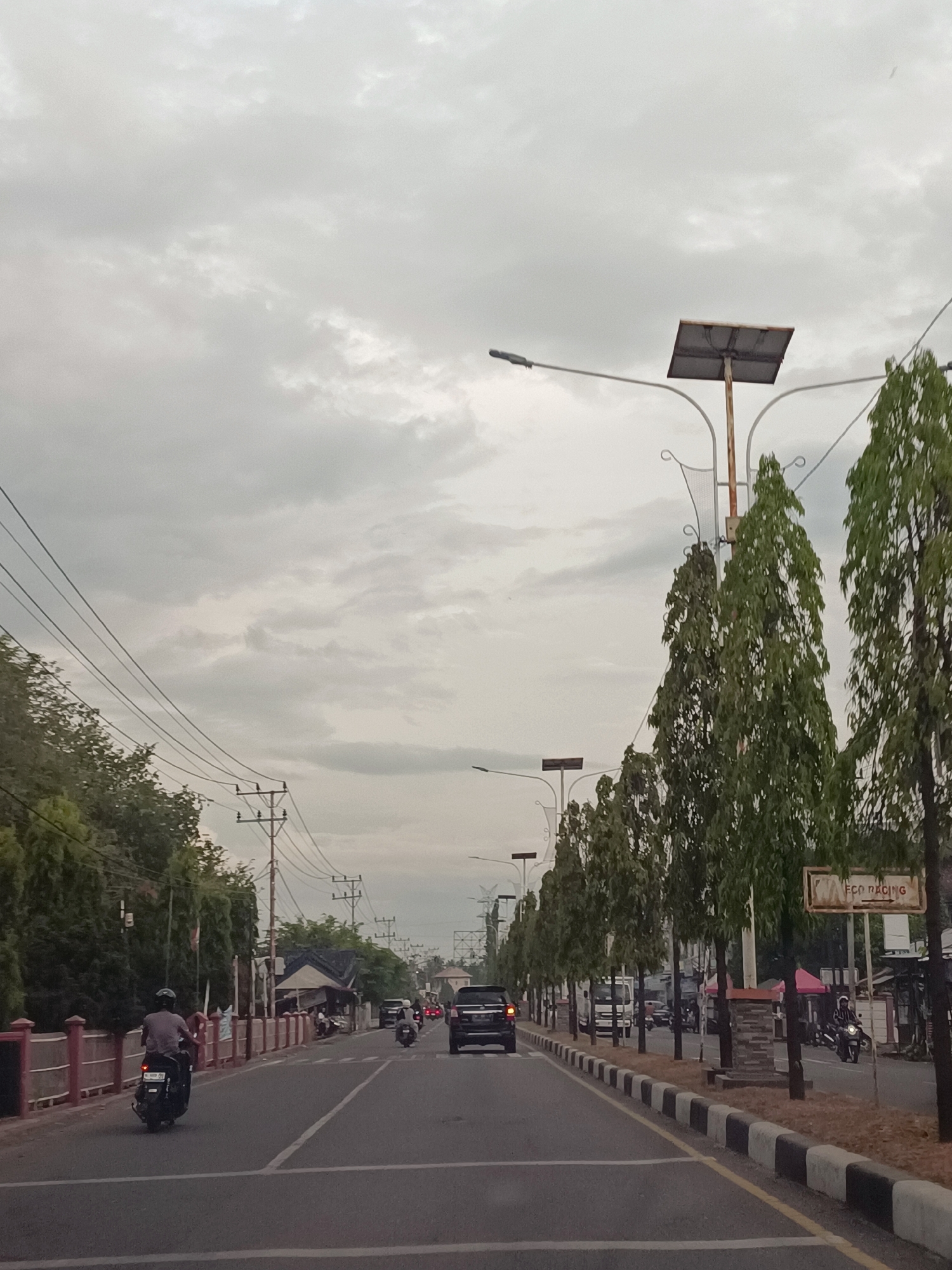 The image depicts a street scene in Paya, Indonesia, under an overcast sky. A paved road with lane markings runs through the center of the frame, flanked by a sidewalk and a line of tall, slender trees on the right. On the left side of the road, a red and white fence runs along the edge, behind which are more trees and a few buildings.
Several vehicles are present on the road. A person on a scooter is riding in the left lane, with a black license plate visible. Further ahead, a black car is traveling in the same lane. In the right lane, there are several other vehicles, including a white truck, a motorcycle with a rider wearing a helmet and jacket, and a black SUV.
Streetlights with solar panels are visible along the roadside, as are power lines and poles. A sign with text that appears to read "ECO RACING" is mounted on a pole near the right edge of the image. The overall impression is of a moderately busy street in a town or city, likely during the daytime given the ambient light, despite the cloudy conditions. The vegetation suggests a tropical or subtropical climate.