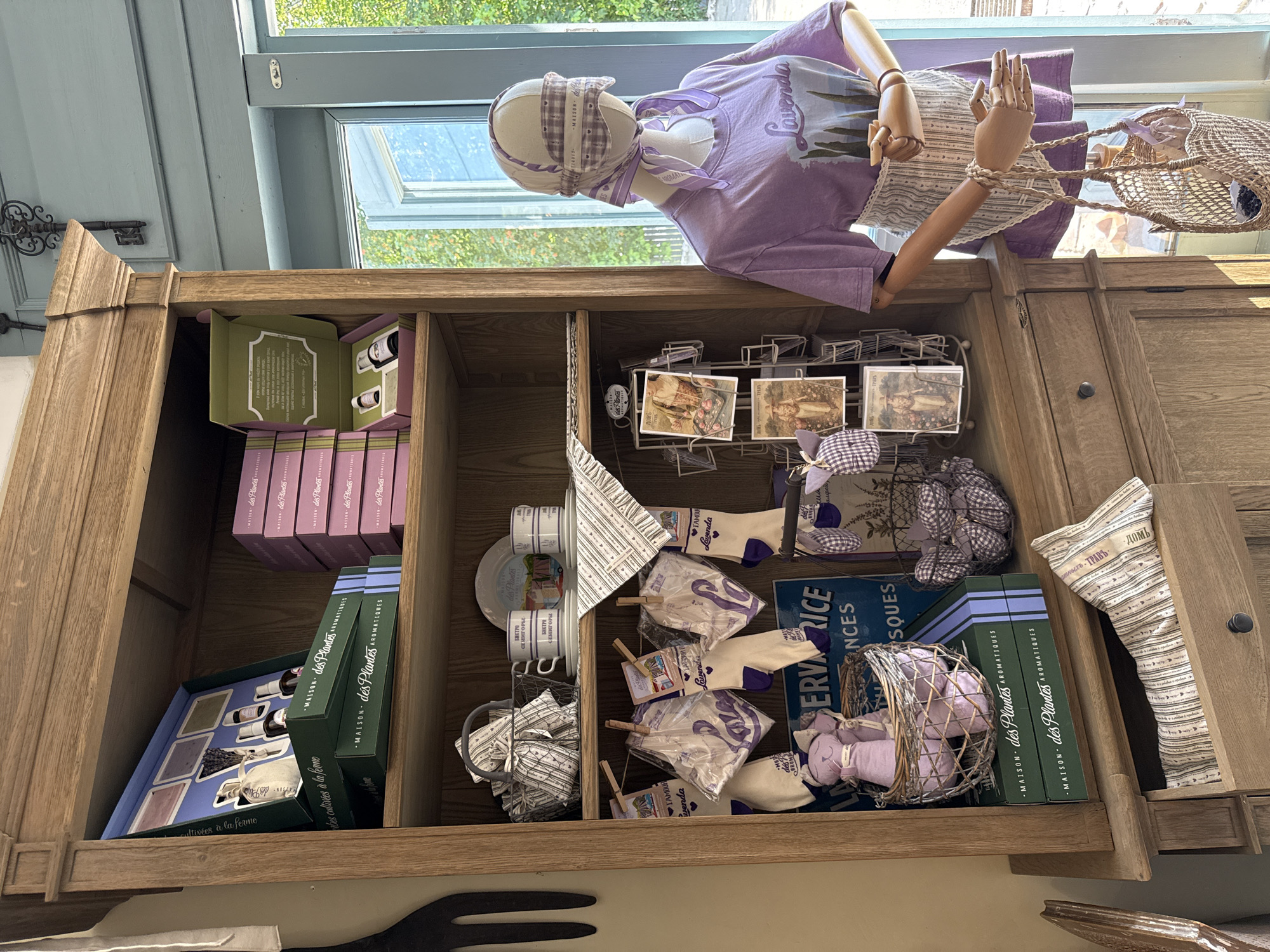 A wooden hutch is filled with various merchandise, including gift boxes, cups, plates, socks, and stuffed animals, all with a lavender theme. In front of the hutch, a mannequin is dressed in a lavender t-shirt and an apron, holding a straw bag. The scene appears to be a retail setting, possibly a shop in Semogorskiy, Russia, judging by the style of the merchandise. The overall impression is one of a charming, rustic store display.