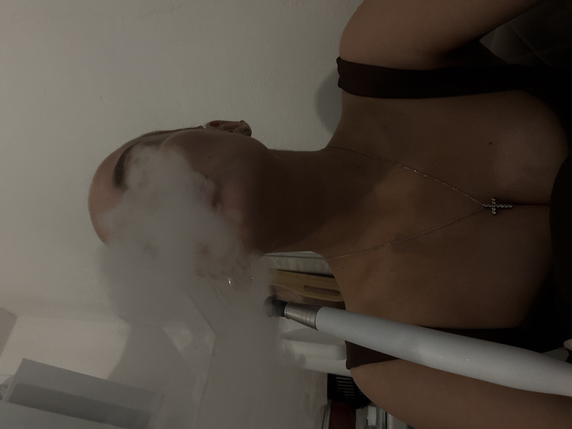 A woman with a shaved head is exhaling a large cloud of vapor from a white vape pen. She is wearing a brown top and a silver necklace with a cross pendant. The location appears to be indoors, possibly a residence. The backdrop includes a white wall and a glimpse of furniture. There is no clear indication of the city of Chaikovsky, Russia.