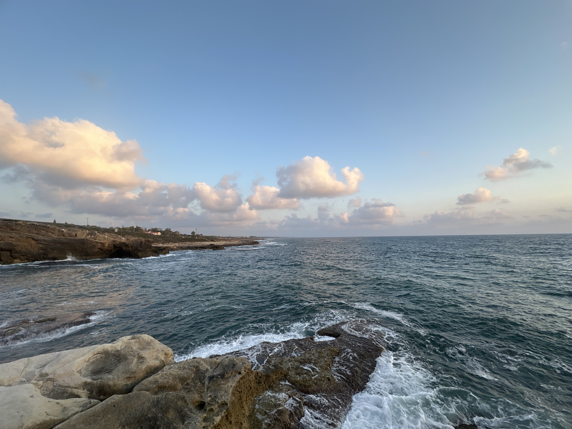 The image depicts a coastline in the Mate Asher Regional Council, Israel. The foreground features rough, textured rocks, some of which are partially submerged by the sea. Waves are actively crashing against these rocks, creating white foam and spray. The sea itself is a deep teal color, with visible ripples and choppiness, indicating a moderate wind.
In the middle ground, the coastline continues with rocky cliffs and a hint of a beach or sandy area further in the distance. Some low-lying buildings and vegetation are visible along the clifftop, suggesting human habitation. The horizon is marked by a clear separation between the sea and the sky.
The sky above is a pale blue, with scattered clouds. Some of these clouds are illuminated by warm, golden light, suggesting it is either early morning or late afternoon, likely during sunrise or sunset. The overall scene conveys a sense of natural beauty and coastal landscape. There are no visible people in the image.