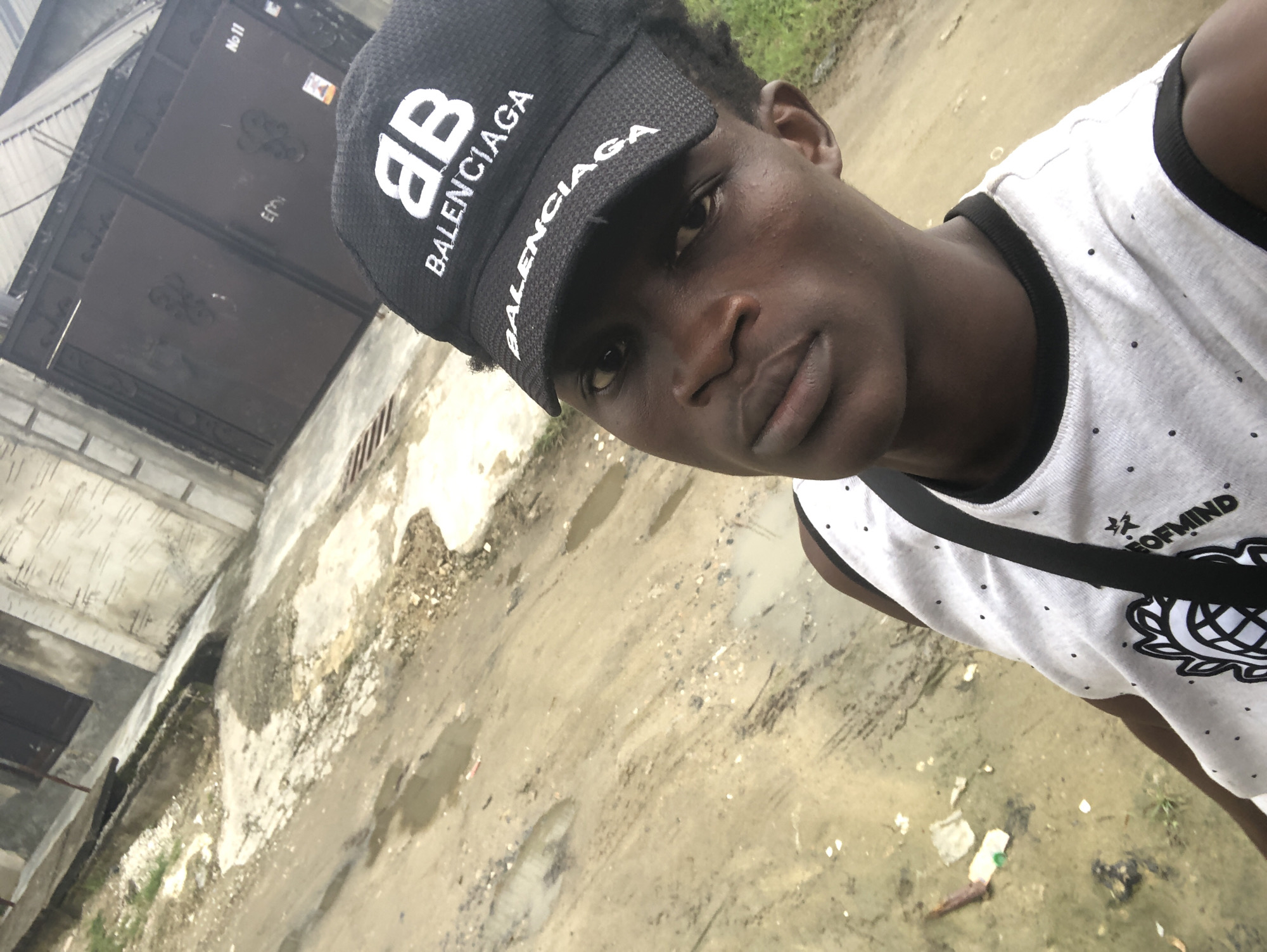 A close-up, slightly overhead selfie shows a young Black male wearing a black Balenciaga baseball cap and a white sleeveless shirt with black polka dots. He is looking directly at the camera with a neutral expression. The shirt has a black trim around the neckline and armholes, and a black graphic design with the words "OF MIND" visible on the front. A black strap is visible across his chest.
The background suggests an outdoor urban environment. The ground is a mixture of dirt, gravel, and puddles of water, indicating recent rain or poor drainage. In the mid-ground, a large dark metal gate with decorative scrollwork is visible, suggesting the entrance to a property or compound. To the left of the gate, a light-colored concrete building with vertical lines is present. A sign with the number "No 11" is attached to the gate. The overall scene depicts a somewhat unkempt urban street or alleyway. The image quality is adequate but not high-resolution. There is no indication of the specific location within Ado, Nigeria.