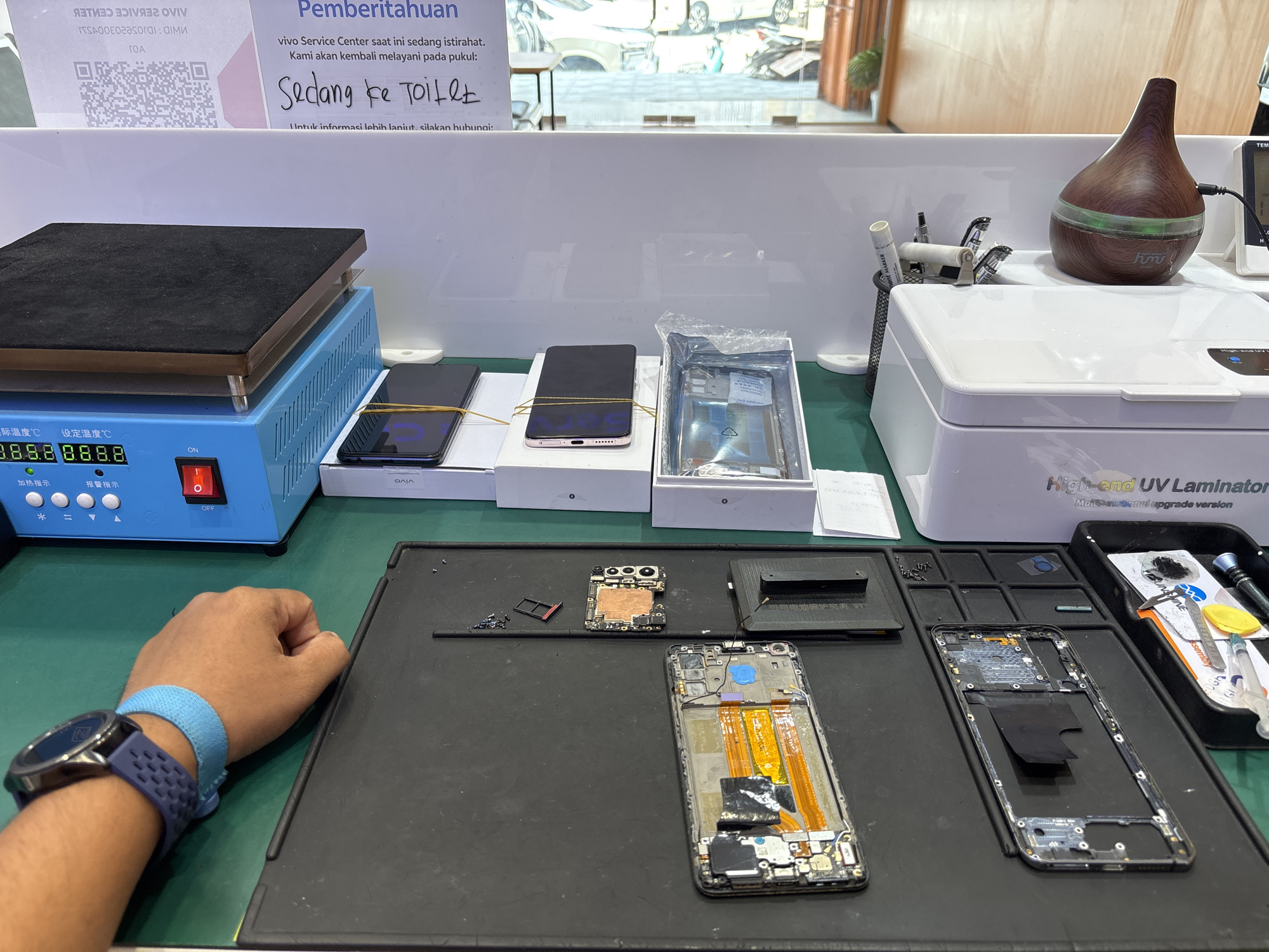 The image depicts a phone repair shop in Lhokseumawe, Indonesia. The scene is set on a workbench with a green mat. In the foreground, a person's left arm is visible, wearing a blue wristband and a dark blue smartwatch. The arm rests on the green mat, positioned near a black tray containing disassembled phone components.
On the workbench, various electronic repair tools and devices are arranged. A blue heating plate with a black surface is prominent on the left. To its right, two mobile phones are laid out, one with its screen facing up and secured with yellow rubber bands, and another on its side. Next to these phones is an open box containing what appears to be a phone component, possibly a battery or screen, wrapped in protective plastic.
Further back on the workbench, a white device labeled "High-end UV Laminator" is visible. To the left of this laminator, a cylindrical wooden-patterned diffuser with a green light emanates from its center is placed. A pen holder with various tools, including pens and tweezers, is situated next to it.
In the background, a sign is displayed that reads "Pemberitahuan" (Announcement) in Indonesian, followed by information about the vivo Service Center being temporarily closed for a break and will resume service at a specified time. Below this, a handwritten note in Indonesian says "Sedang ke Toilet" (Going to the toilet). A QR code is also visible on a white surface in the background. The environment suggests an indoor setting, possibly a commercial space, with hints of reflections of the outside world in the background windows.