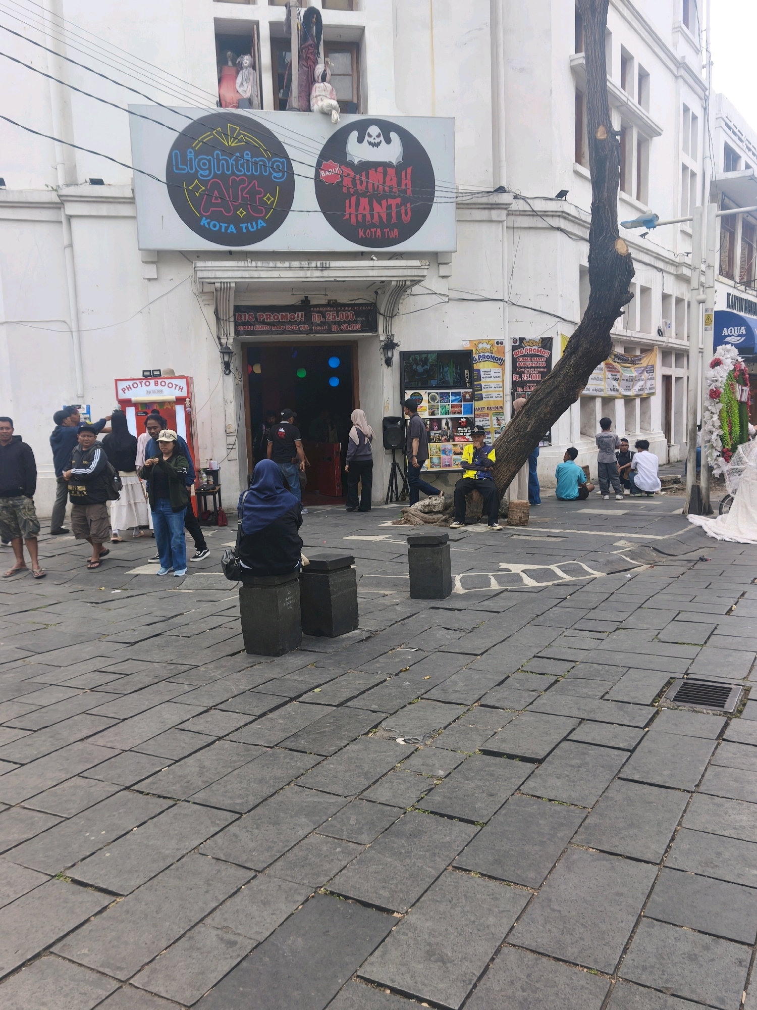 This image captures a street scene in Kota Tua, West Jakarta, Indonesia. In the foreground, a gray cobblestone plaza is visible, with some areas showing slight wear and cracks, and small patches of grass growing through. Two dark gray cube-shaped planters are present on the plaza.
In the midground, a white building dominates the scene. Two large circular signs are affixed to the building's facade. The sign on the left reads "Lighting Art KOTA TUA" in colorful lettering. The sign on the right features a cartoon ghost graphic and the text "RUMAH HANTU KOTA TUA." Below these signs, there is an entrance to a building with a large banner proclaiming "BIG PROMO!!" with text indicating pricing for "RUMAH HANTU" and "TOULER". The entrance appears to be a doorway with red accents and colorful lights visible inside.
Several individuals are present in the scene. On the left side of the image, a group of people are gathered, some appearing to be posing or interacting. A red "PHOTO BOOTH" structure is also situated on this side. To the right of the building's entrance, a tree with a prominent, gnarled trunk leans into the scene, its bark rough and textured. A group of young men are seated on the ground near the base of this tree. Further to the right, another entrance to a building is visible, with a banner that appears to be for an "AQUA" product, and further decorations including white fabric and floral arrangements.
The overall environment suggests a public space, likely a tourist or entertainment area within the historic district of Kota Tua, West Jakarta, given the architectural style of the building and the presence of attractions like a "haunted house" and a photo booth. The lighting indicates it is daytime, with shadows cast by the tree and some of the people.