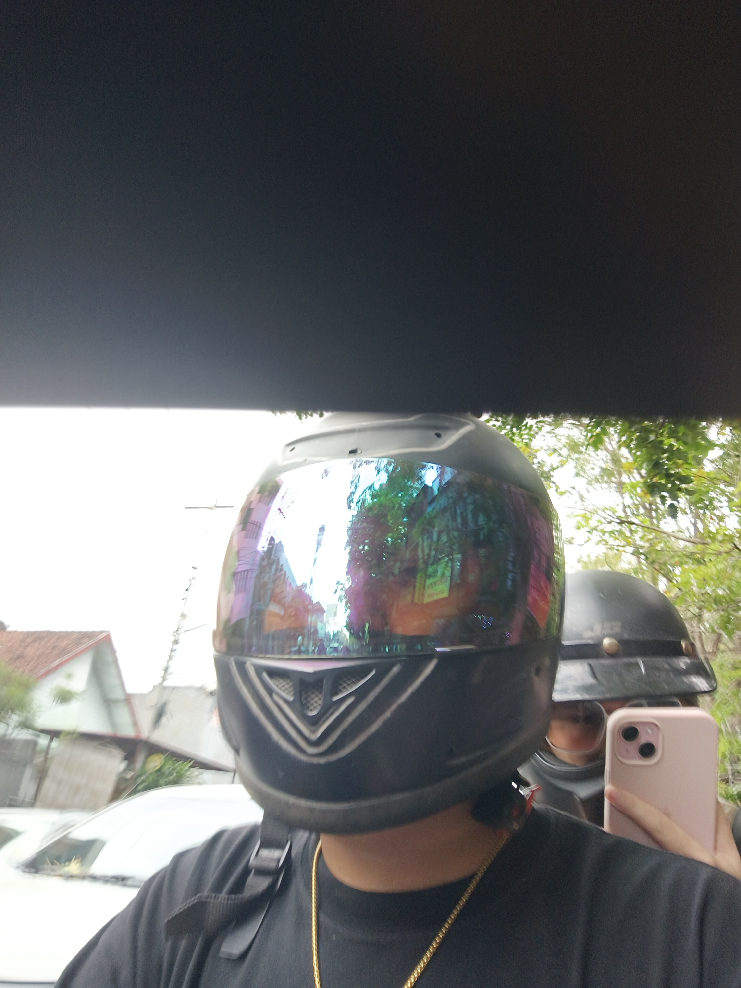 The image shows a selfie taken in Denpasar, Indonesia. The primary subject is wearing a black motorcycle helmet with a reflective visor that shows a distorted reflection of buildings and trees. A person in the background is also wearing a black motorcycle helmet. The person in the foreground is holding a pink smartphone to take the selfie, and a gold chain necklace is visible. In the background, there are houses with terracotta roofs and lush green trees, suggesting a semi-urban or suburban environment within Denpasar. The sky is bright and overcast, and there is a white car in the lower left portion of the frame. The image appears to be taken outdoors, possibly on a street or driveway in Denpasar.