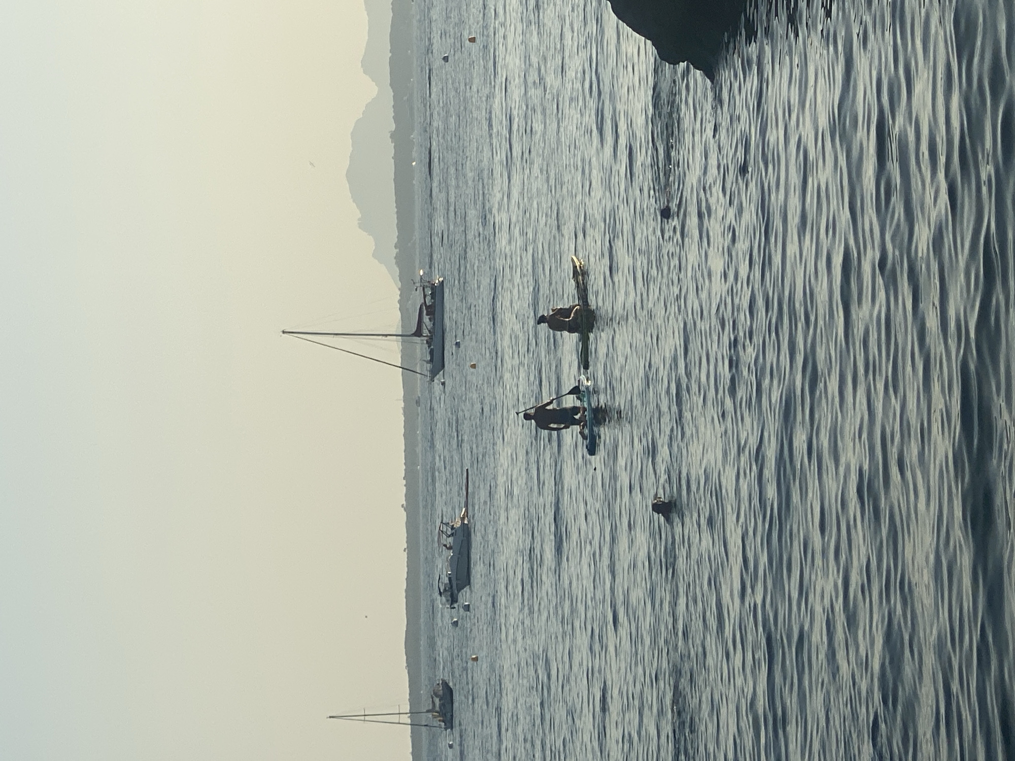 The image shows a wide view of the sea off the coast of Antibes, France, with a hazy, pale sky. In the foreground, the water is choppy and a dark rock juts out from the right side. There are two people on paddleboards in the middle ground, one kneeling and paddling, the other sitting. A dog is swimming in the water between them. In the background, several sailboats and a motorboat are anchored or moving slowly, and a distant, mountainous coastline is visible on the horizon. The scene appears to be a tranquil summer day.