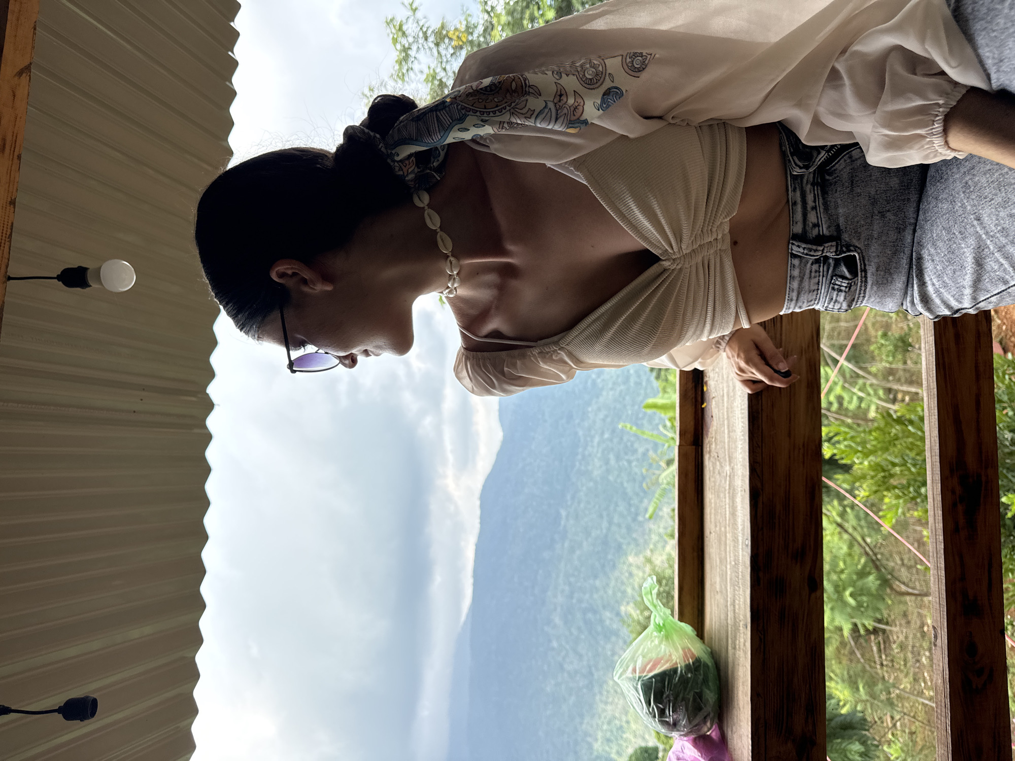 A young woman with dark hair pulled back and a printed scarf tied to it, wearing sunglasses, a white shell necklace, a white ruched crop top, and acid-wash jeans, is seated at a wooden table outdoors. She is looking to her left, away from the camera. A plastic-wrapped bundle of what appears to be produce sits on the table to her left. In the background, lush green mountains under a cloudy sky are visible. A light fixture hangs from the corrugated roof above. The scene is likely set in Son Dien, Vietnam, judging by the mountainous terrain and vegetation.