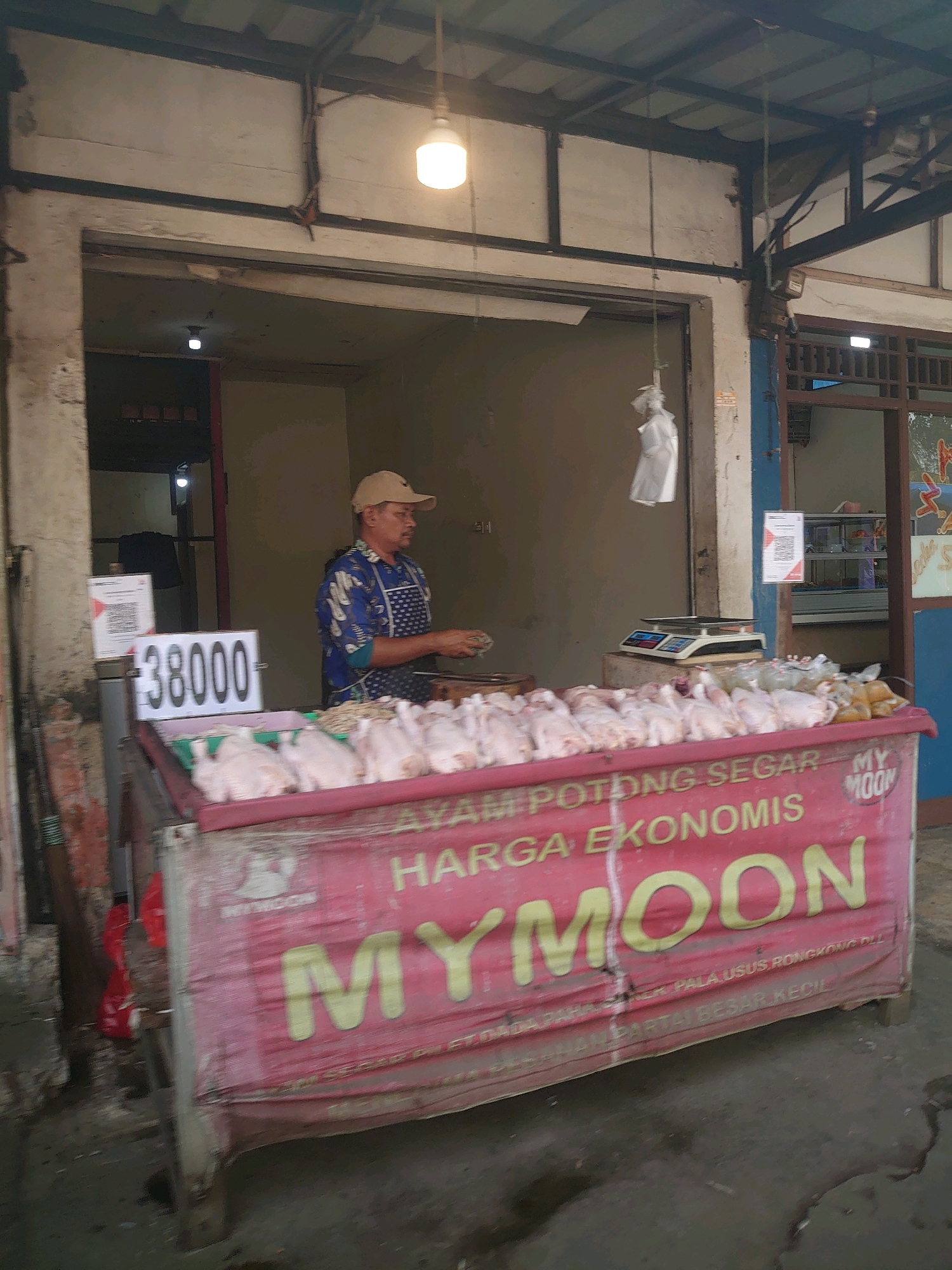 The image depicts a street market scene in Depok, Indonesia, focusing on a vendor selling fresh chickens. A man wearing a tan baseball cap and a blue shirt with a patterned apron stands behind a stall, arranging chickens. The stall is adorned with a large red banner displaying text in Indonesian. Prominently featured on the banner is the brand "MYMOON" in yellow lettering, along with phrases like "AYAM POTONG SEGAR" (Fresh Cut Chicken) and "HARGA EKONOMIS" (Economical Price). To the left of the vendor, a price tag displays "38000" next to a QR code. The background reveals a simple, open-air market structure with a tin roof and exposed beams. Several raw chickens are laid out on the stall, and a digital scale is visible in the background. The ground appears to be asphalt, with some damp patches. The overall impression is that of a typical local food market.