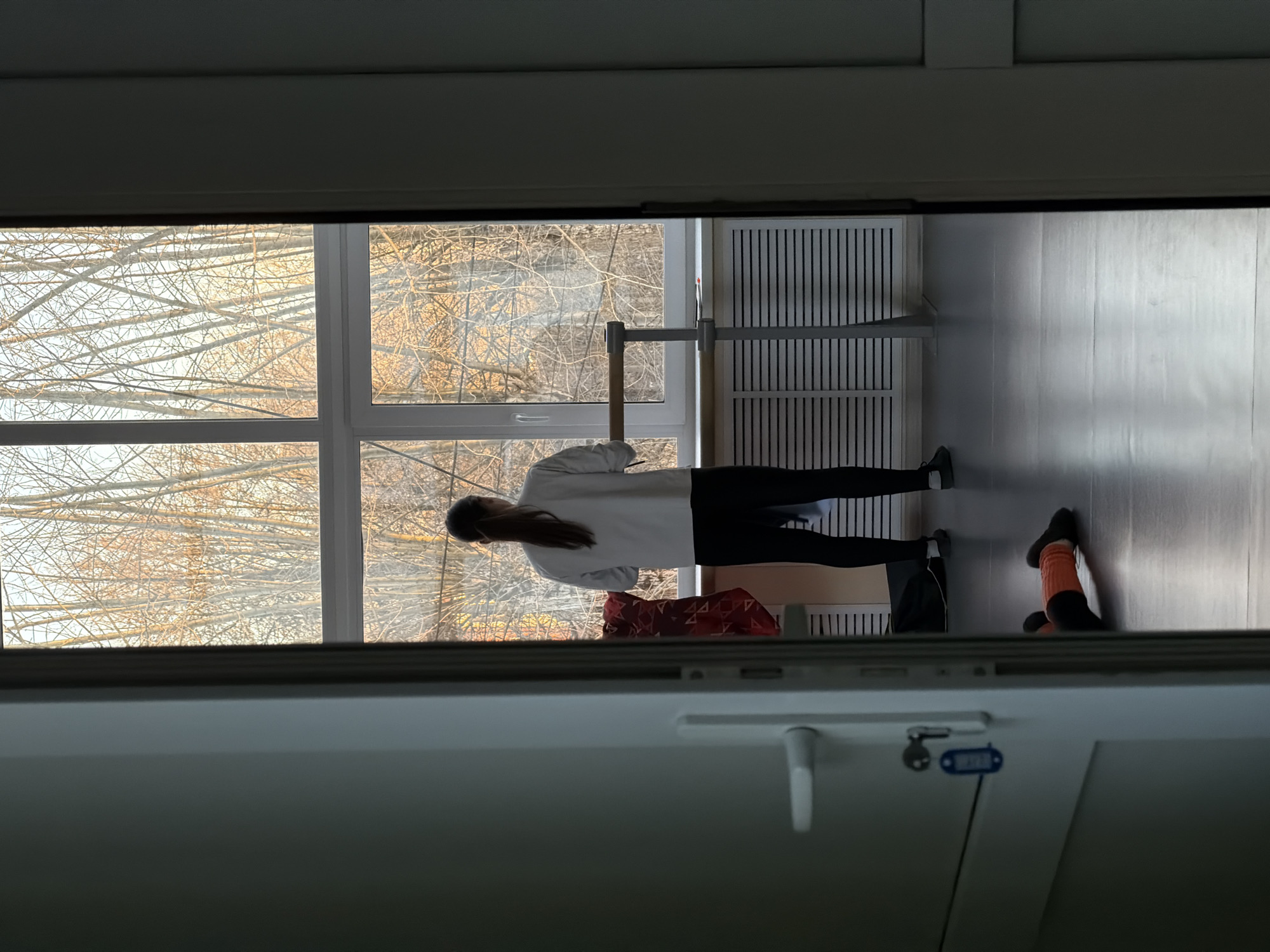 A medium shot, taken from inside a room looking through an open window at a woman standing in front of a barre. The woman is wearing black pants and a white long-sleeved shirt. She is facing away from the window, toward the room. Behind the woman, there is a window with a view of bare trees. To the right of the woman, there is a barre and a vent on the wall. There are personal items in a pile in the corner on the floor. The floor is made of wood. The shot is taken from Perm, Russia.