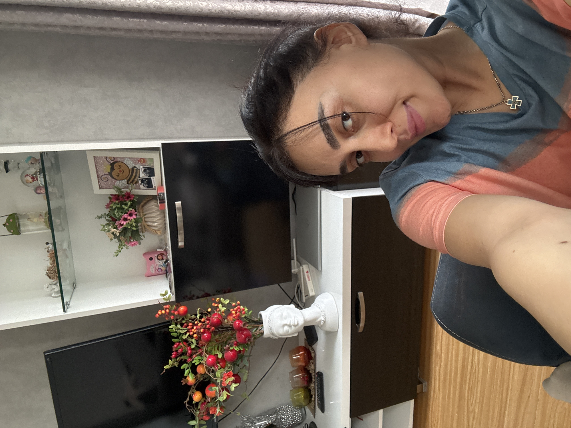 The image, currently oriented vertically (rotated 90 degrees to the left from its original capture), is an indoor selfie featuring a woman and a section of a living room. The woman has dark hair pulled back and looks towards the camera with a slight smile and tilted head. She wears a blue and orange/pink tie-dye t-shirt and a silver chain necklace with a black and silver cross pendant. Part of her arm and her right leg are visible, suggesting she is seated.
The background reveals a modern white and dark-colored TV stand or cabinet, characteristic of a residential interior in Tbilisi, Georgia. Its open shelves on the left display various decorative items, including small figurines, a snow globe, a framed picture of a cartoon bee, a small pink ornament, and an artificial pink floral arrangement. A black flat-screen television is partially visible below these shelves. On the cabinet's surface, a white ceramic vase shaped like a classical head holds an arrangement of artificial red berries, green leaves, and small red/orange fruits. Three small, translucent colored containers (amber, red, green) and a remote control are also on the stand. The upper background shows a light gray textured wall or curtain, and the floor is light-colored wood laminate. The scene conveys a relaxed, casual moment captured during the daytime; no discernible text is visible on any objects.