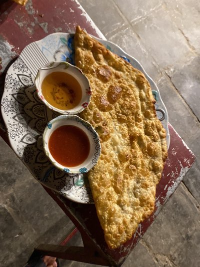 The image shows a fried pastry, likely bolani, served on a patterned plate with two small bowls of dipping sauces. One sauce is a clear amber color, possibly honey or a sweet syrup. The other sauce is a red color, likely a chili sauce. The plate sits on a weathered red wooden surface, possibly a low stool or table. The location is likely Kandahar, Afghanistan, based on the presence of bolani, a common Afghan dish. The lighting suggests the photo was taken indoors or in a shaded outdoor area. No people are visible, though a foot appears in the lower left corner.