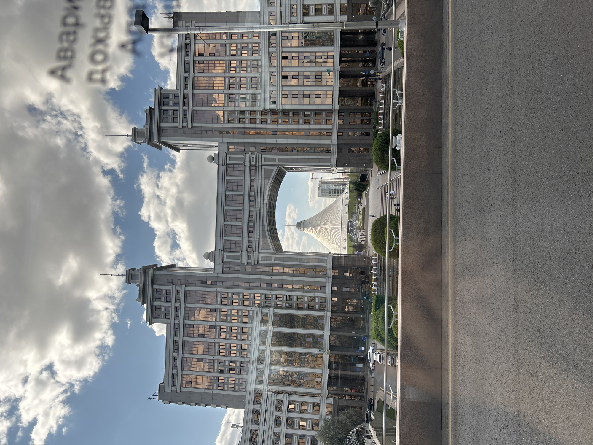 The image captures a wide view of a modern city square in Astana, Kazakhstan, featuring the iconic Bayterek Tower in the distance, visible through a large archway formed by two symmetrical, multi-story buildings. The archway itself is part of a grand, contemporary architectural complex. The two buildings flanking the arch are similar in design, with a grey facade and numerous windows reflecting the sky and clouds. The sky is partly cloudy, with patches of blue visible.
In the foreground, a section of an asphalt road is visible, bordered by a low, dark brown wall. Beyond the wall, there is a landscaped area with green bushes and trees, as well as fountains. Several people are seen walking in the square, and a few cars are parked or driving in the background. The overall scene suggests a public space in a modern urban environment, likely during daylight hours. Some text, appearing to be in Russian, is superimposed on the sky in the upper right corner of the image.