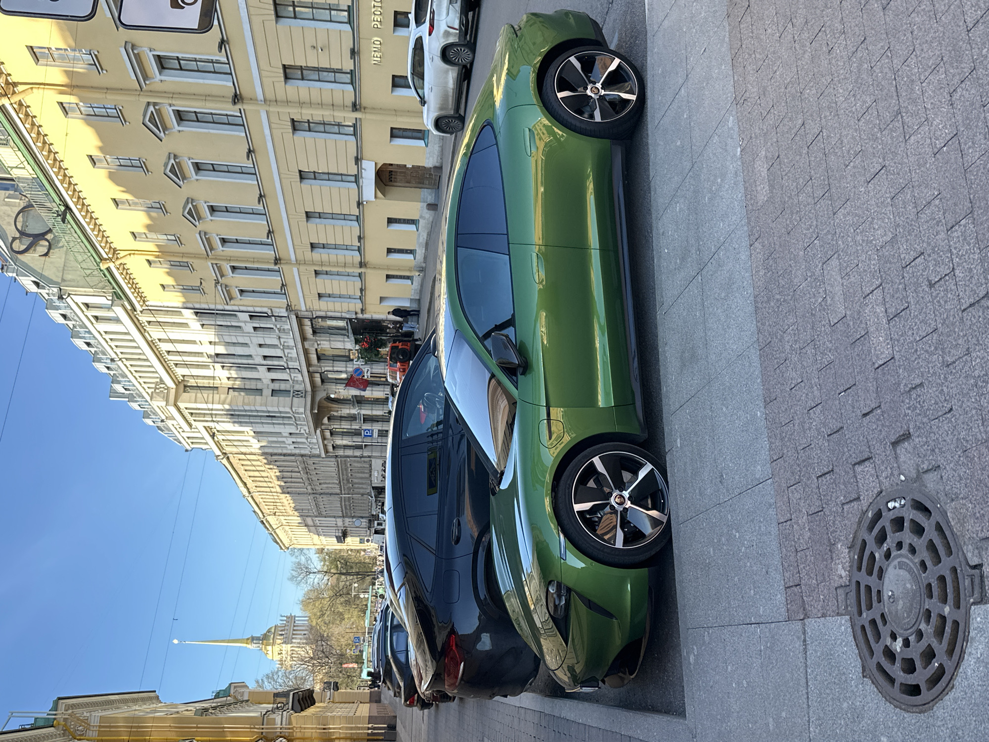 The image, presented rotated 90 degrees clockwise, depicts a sunlit city street scene in Saint Petersburg, Russia. The clear blue sky overhead suggests a bright day, likely late morning or early afternoon, given the shadows cast by the buildings.
In the immediate foreground, a striking lime-green modern sports car, possibly a Porsche Taycan given its distinctive design and wheels, is parked along a grey paved sidewalk. Its black and silver multi-spoke alloy wheels are a prominent feature. Behind it, a dark-colored SUV or sedan is parked, with a line of other vehicles extending down the street. A circular metal manhole cover with a patterned grate is visible on the sidewalk.
The street is flanked by tall, ornate historical buildings, characteristic of European architecture. On the left side, a grand, light yellow building with numerous windows, intricate cornices, and decorative window frames dominates the view. Buildings on the right side appear lighter, possibly white or light grey.
In the background, a very distinct golden spire with a pointed top can be seen rising above the rooftops, strongly identifying the location as Saint Petersburg due to its resemblance to the Admiralty Building. Some bare trees are visible, hinting at late winter or early spring.
Visible text on a sign on the yellow building reads "NEMO PЕСТО," where "РЕСТО" likely abbreviates "ресторан" (restaurant) in Russian. The scene is static, capturing the elegant urban landscape with its historical architecture and modern elements.