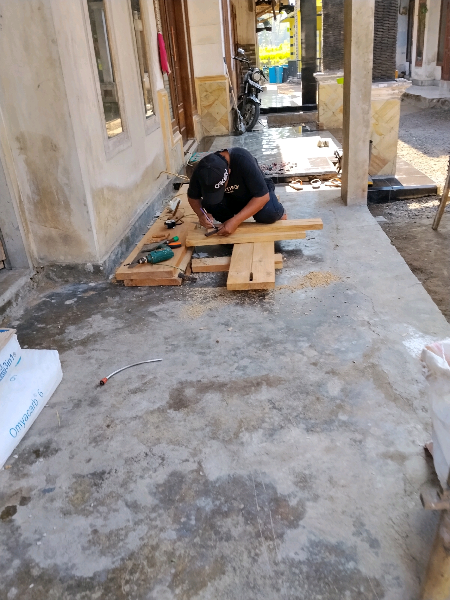 A male, wearing a black t-shirt and dark pants, kneels on a concrete ground in Panggungan, Indonesia, working on wooden planks. He is holding a pencil in his right hand and appears to be marking a measurement on the wood. A green power drill is placed next to the wood, along with other tools and sawdust. The scene is outdoors, with a building visible in the background, featuring windows and wooden doors. Sunlight casts shadows on the ground. A white bag with blue lettering, possibly containing construction materials, is on the left side of the image. A red and white hose lies on the concrete.