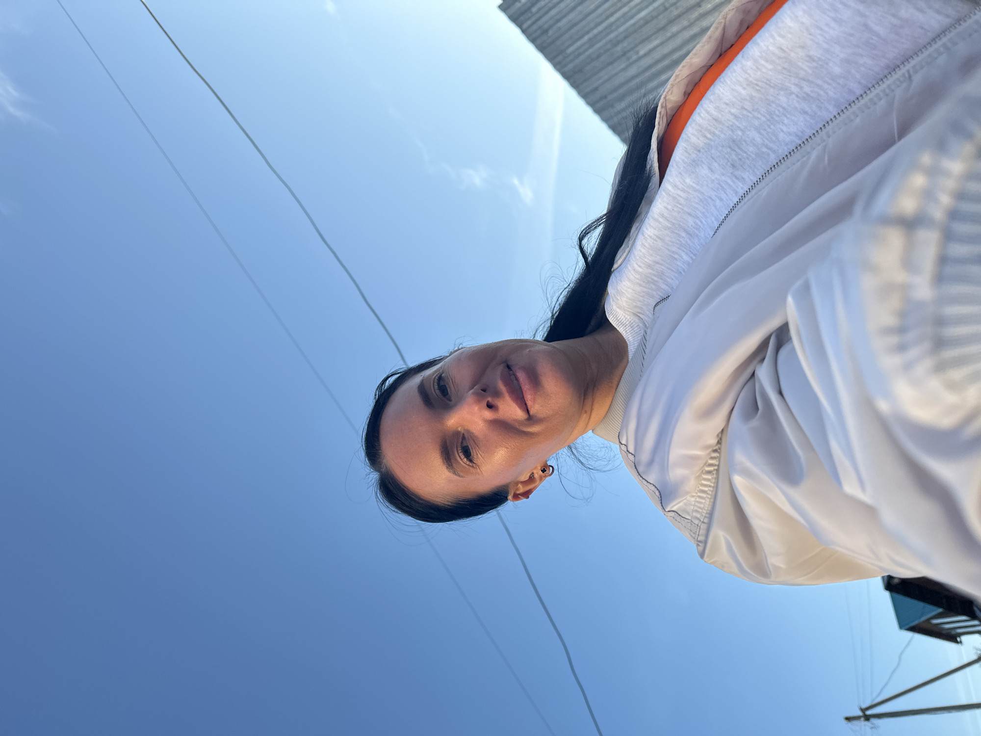 An upward-angled outdoor portrait shows a woman against a clear blue sky. The image was originally captured with a portrait orientation, then rotated 90 degrees counter-clockwise. The woman, with dark hair pulled back, looks towards the camera with a neutral to slightly smiling expression. She is wearing a light-colored jacket over a grey hooded top, with an orange layer visible beneath. The background is dominated by a bright, clear blue sky, with a few faint wisps of clouds. Two thin wires, likely utility lines, cross the sky diagonally. In the upper right corner (when correctly oriented), part of a building with a grey, possibly corrugated, roof is visible. A dark, angular structure is also partially seen in the lower right. The bright and even lighting suggests it is daytime. The setting appears to be an outdoor, possibly urban or suburban, environment in Ольховка, Russia. No text is visible in the image.