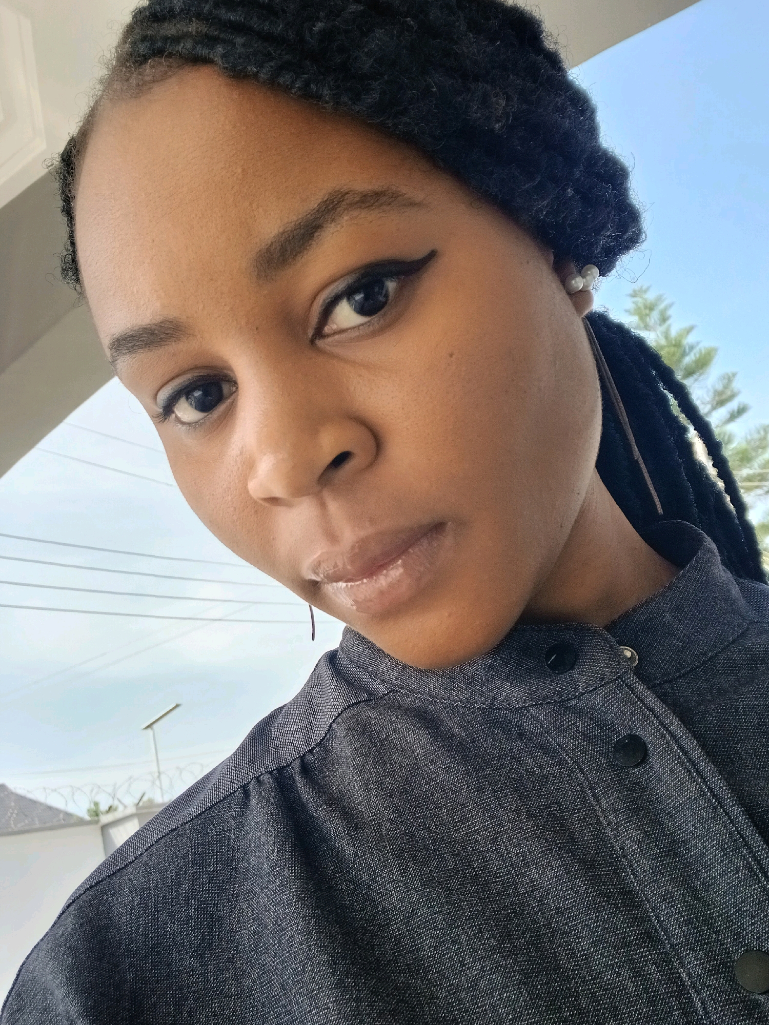 A close-up, slightly tilted portrait shows a young Black woman with braided hair and pearl earrings. She is wearing a dark grey, button-up collared shirt. The background includes a bright blue sky with faint white clouds, the top of a green tree, some power lines, and a sliver of a white building with barbed wire fencing. The scene appears to be outdoors, possibly in a residential area of Gura Top, Nigeria.