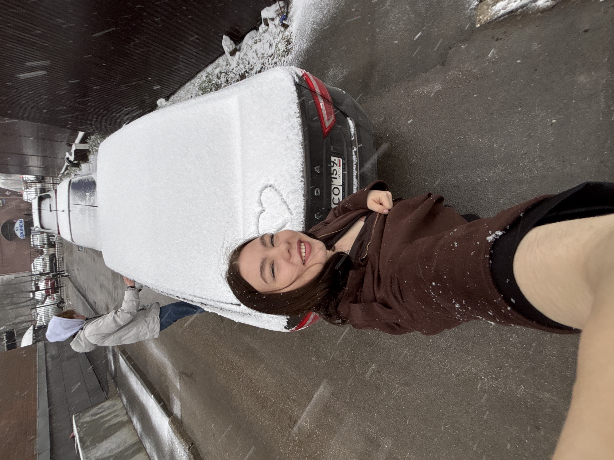 A young woman smiles in a selfie-style photo in Perm, Russia. Snow covers a parked dark-colored car with a license plate reading "CO 159 RUS." A heart is drawn in the snow on the car's trunk. Another person stands near the front of the vehicle, wearing jeans and a light-colored jacket. The street and nearby buildings are visible in the background. It is snowing.
