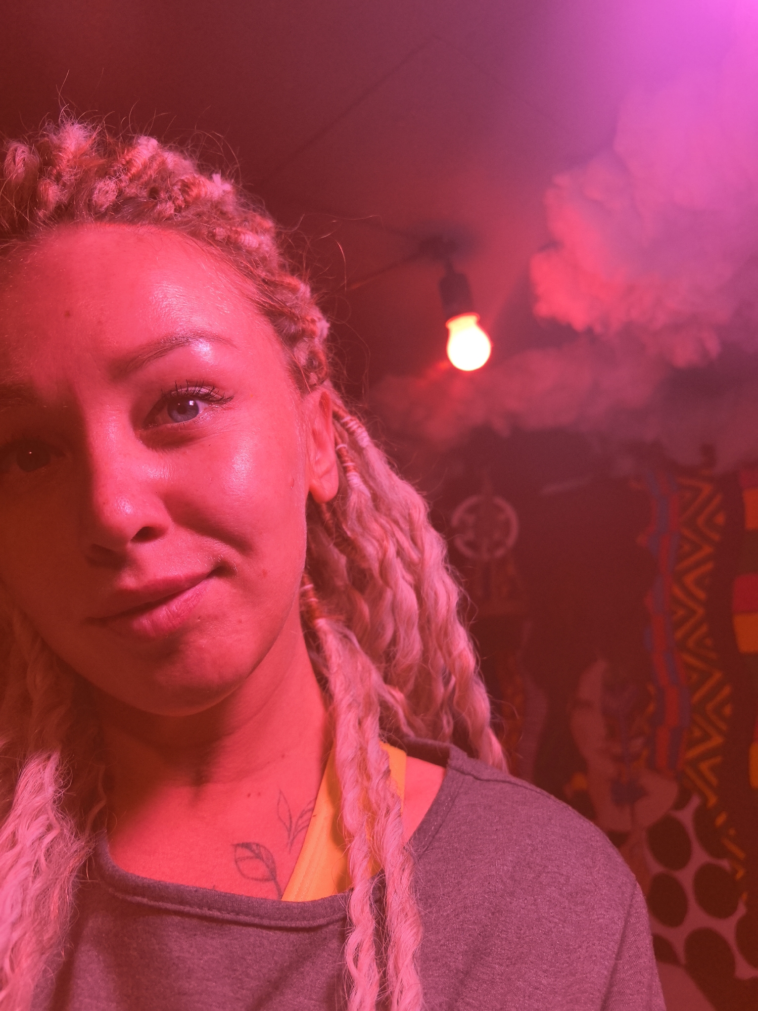 The image presents a close-up, slightly upward-angled portrait of a woman, heavily illuminated by vibrant red and pink lighting. She has fair skin, blue eyes, and extensive light-colored dreadlocks or textured braids that cascade around her. A small, subtle smile plays on her lips, and her gaze is directed towards the camera. On her left collarbone, a tattoo resembling two leaves or a delicate branch is visible. She is dressed in a grey top, layered over a bright yellow garment visible at her neckline.
The setting appears to be an indoor space, possibly a club, themed room, or social venue, characterized by its dim overall lighting dominated by the intense red and pink hues. In the background, a single, bare incandescent light bulb hangs from the ceiling, emitting a distinct white-yellow glow that stands out against the red environment. Above, a cloud-like decorative element is present on the ceiling. The wall behind her is adorned with various dark-toned, abstract patterns, including zig-zags, circular dots, and stylized figures, suggesting a bohemian or artistic aesthetic. Another circular decorative object is faintly visible in the mid-ground.
The scene conveys a relaxed, casual moment, likely a selfie given the close proximity and angle. The predominant artificial lighting suggests nighttime or an interior designed for specific ambiance, rather than daylight. The woman's expression is calm and approachable. No visible text is present in the image. The location is Noviy, Russia, though the image itself contains no explicit geographical markers.
