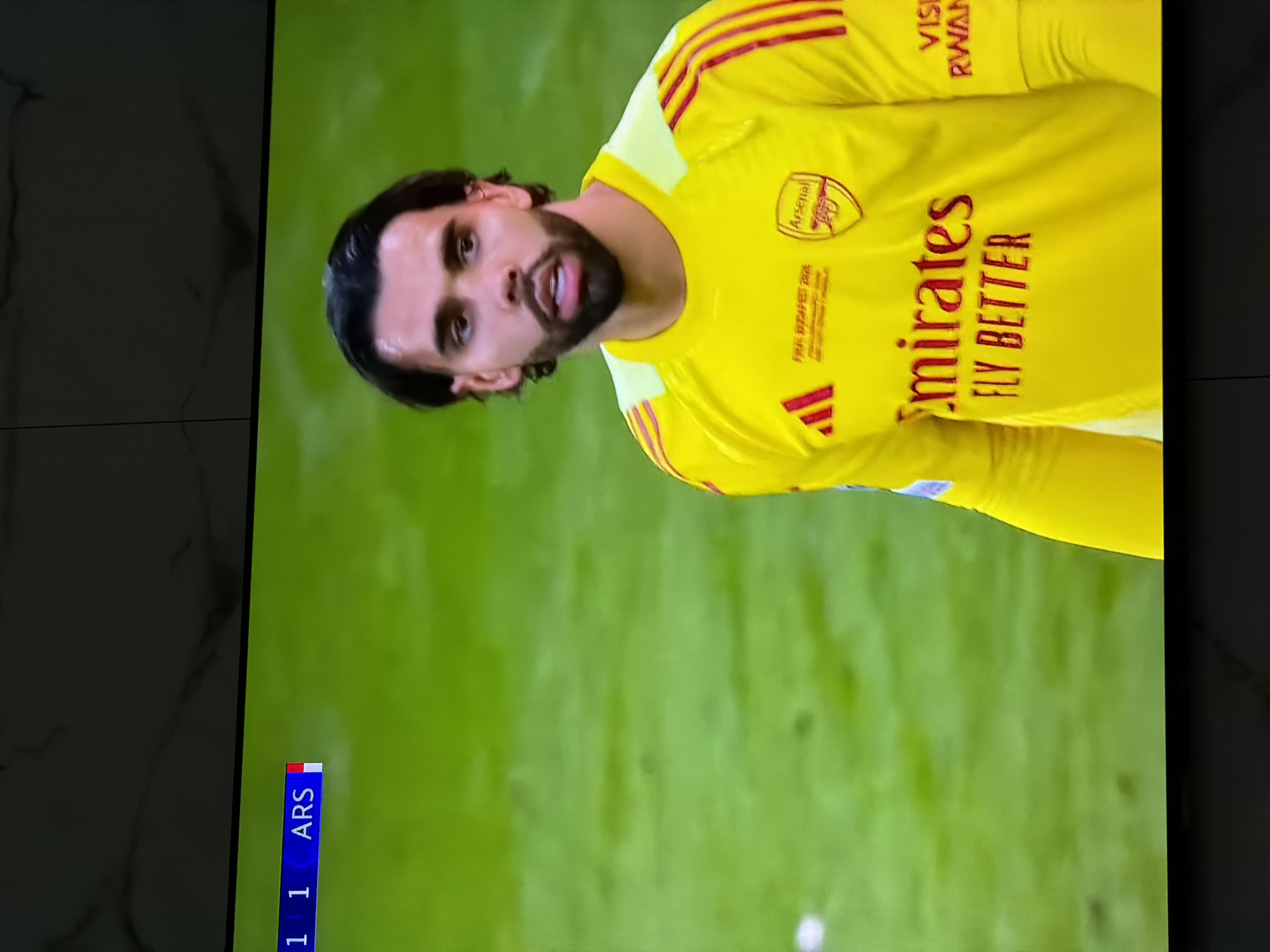 The image shows a man wearing a yellow long-sleeved shirt with the Arsenal logo. The shirt also has the "Emirates Fly Better" advertisement. The text "Visit Rwanda" is visible on the sleeve. The scoreboard displays "1-1 ARS" indicating a soccer match score. The background is a grassy field. The image provides no visual information to determine the location in Asenovgrad, Bulgaria, or whether it is even located there.