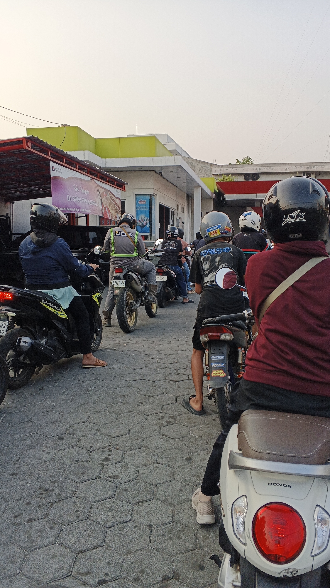 In the city of Kasihan, Indonesia, a group of individuals on motorcycles are queuing at a gas station. The scene is captured from a low angle, looking forward. In the foreground, a person wearing a maroon jacket and black pants sits on a white Honda scooter, facing away from the camera. Further ahead, several other individuals on motorcycles are also waiting. One person wears a reflective green vest over a grey shirt and black pants. Another is on a black motorcycle, wearing a black t-shirt with a graphic on the back and black shorts. The gas station features a prominent yellow structure and a canopy with a red roof. The ground is paved with hexagonal grey interlocking stones, and there are a few oil stains visible. The sky is overcast with a pale yellow hue, suggesting either early morning or late afternoon light.