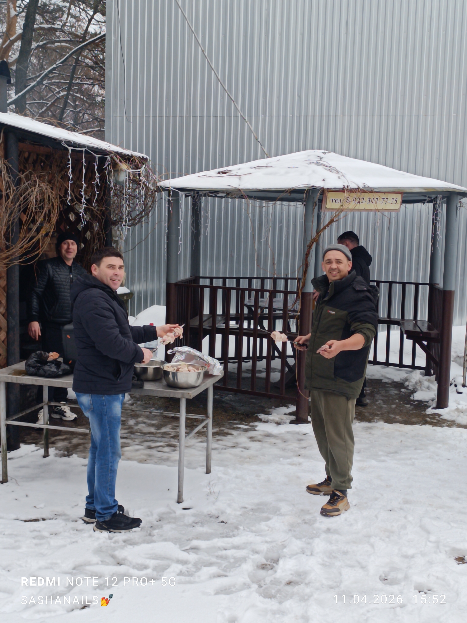 In Chaikovsky, Russia, on April 11, 2026, at 15:52, four men are preparing food outdoors in a snowy environment near two shelters. One shelter is adorned with vines and lights, the other is a gazebo-like structure with a sign displaying a phone number. Two men are actively involved in preparing what appears to be meat skewers; one is putting meat onto skewers from a metal bowl on a metal table, and the other is holding a skewer with meat. The other two men stand by or behind the structures. The background features a corrugated metal wall.