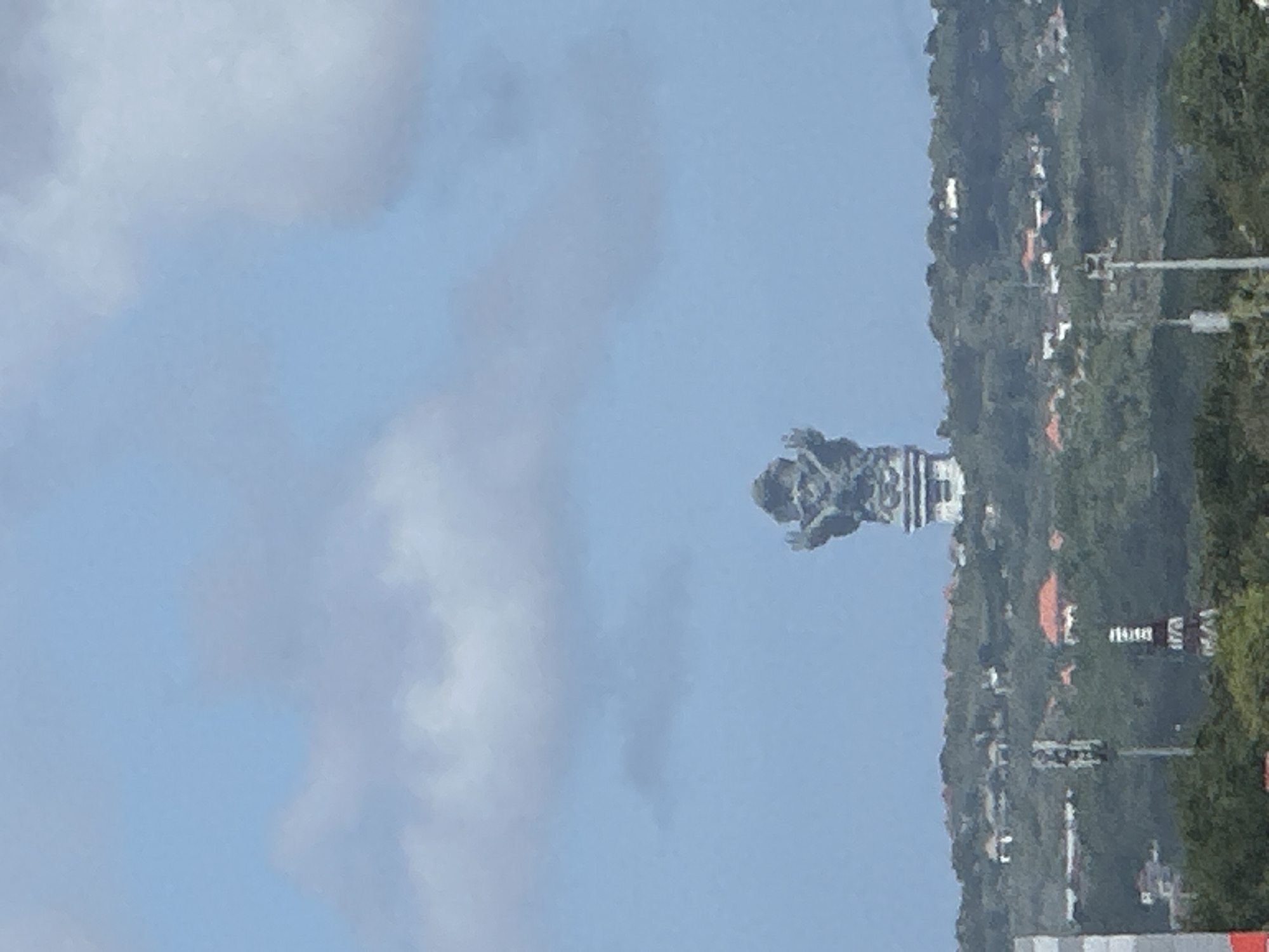 The image shows a large, grey, stone statue of Vishnu riding on the Garuda, located in Ungasan, Kuta South, Badung Regency, Bali, Indonesia. The statue is in the middle ground and stands against a sky with light clouds. To the right, the image shows a hillside with trees and some buildings, including towers and red-roofed structures.