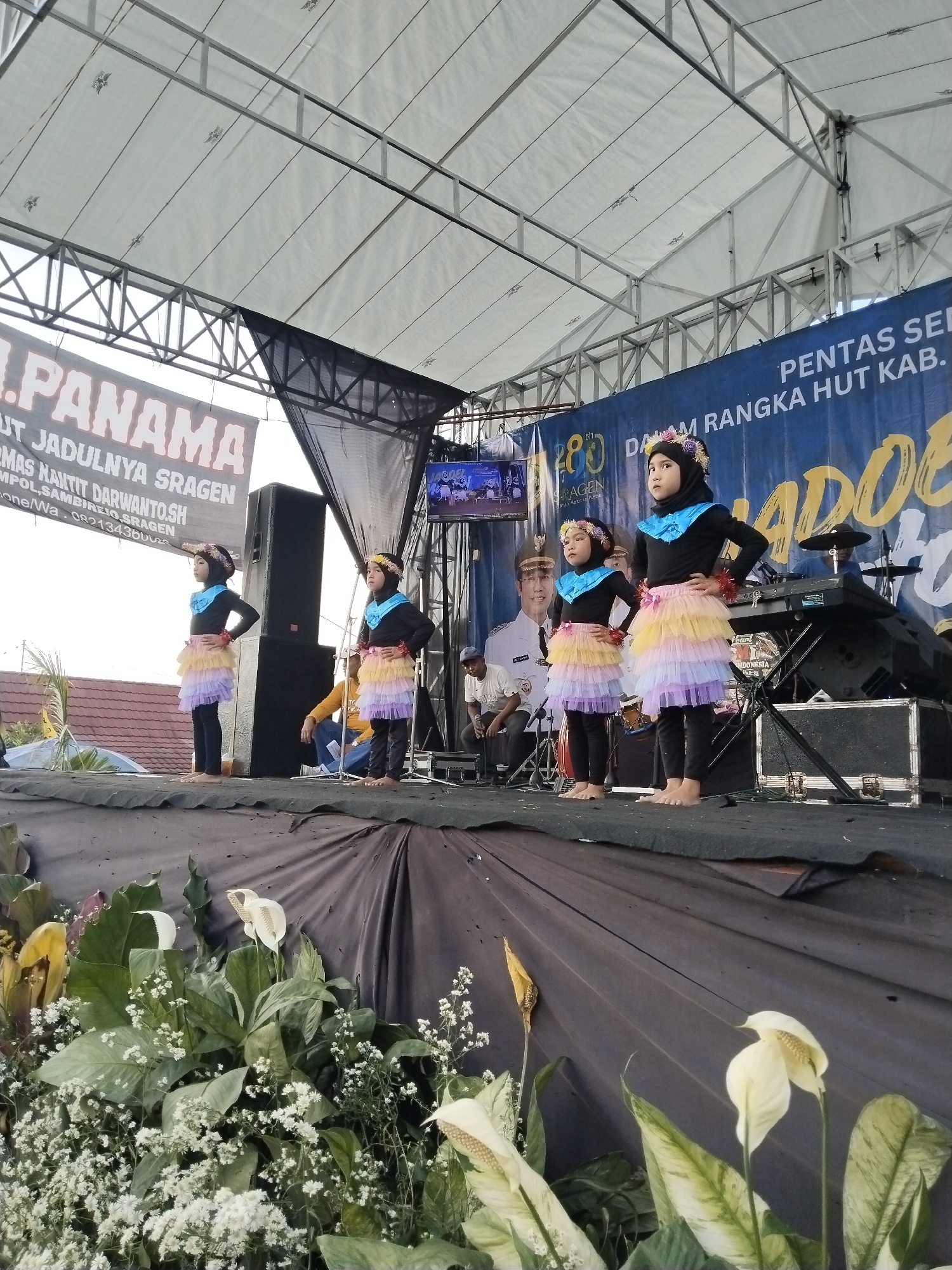 The image captures a scene on a stage, potentially located in Sambirejo, Indonesia. Four young girls stand in a line, each wearing a black top, black leggings, and a multi-layered tutu skirt made of light purple, yellow, and orange tulle. Each has a headpiece or floral crown.
Behind them, on the stage, there are several adults. One man in a white shirt and possibly a formal hat sits on a chair. To the right is a keyboard and a drum set. A television screen hangs above the stage, displaying images.
Banners surround the stage. One banner reads "PENTAS SENI DALAM RANGKA HUT KAB." Another banner reads "I. PANAMA UT JADULNYA SRAGEN" and includes the name "H. TOMAS HANTIT DARWANTO.SH."
The front of the stage is decorated with white and cream-colored flowers, and green foliage. The stage is draped with a black cloth. The event seems to be an outdoor performance or celebration under a canopy roof.