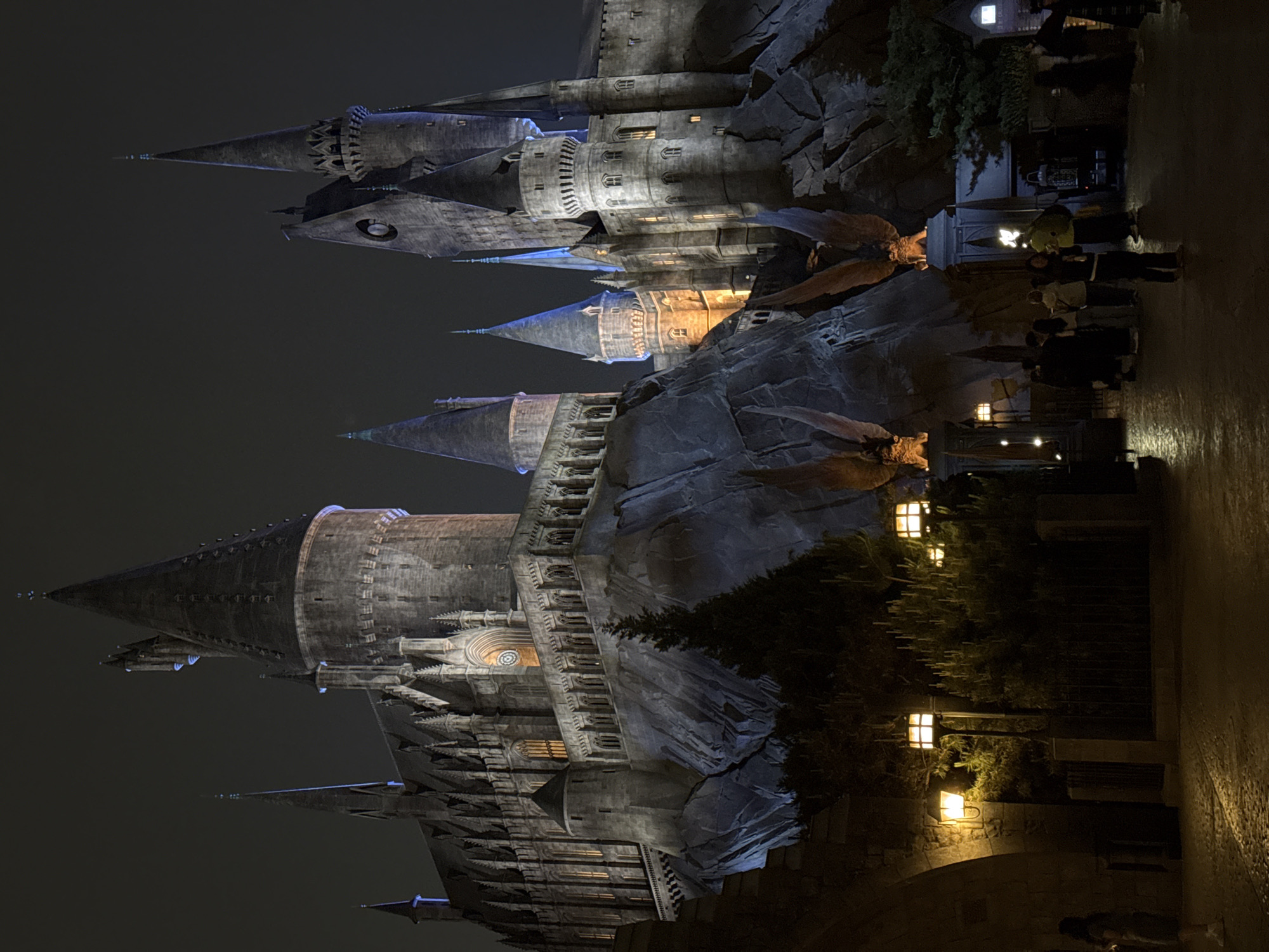 The image shows a nighttime view of Hogwarts Castle at Universal Studios. The castle is constructed against a rocky landscape. There are numerous towers and spires, illuminated with artificial light. In the foreground, there's a pathway with streetlights. Groups of people are present along the pathway. The location is not in Tongzhou District, China, but in a theme park.