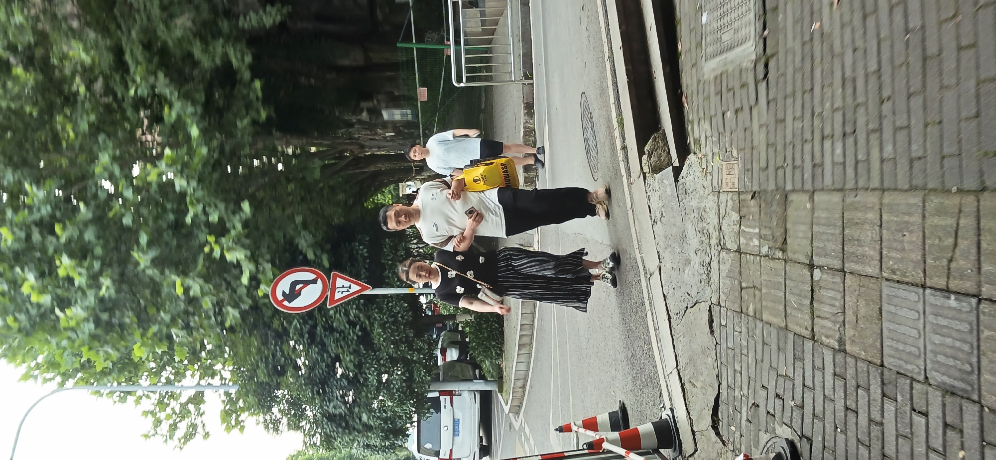 A street scene in Jiulongpo District, China, shows three individuals walking along a sidewalk next to a road. The foreground contains traffic cones and a barrier. A white vehicle is partially visible. A road sign with a "no U-turn" symbol and a Chinese character is present. The sidewalk is paved with rectangular blocks. One person carries a yellow object labeled "LOWASP." The background includes trees and a metal railing.